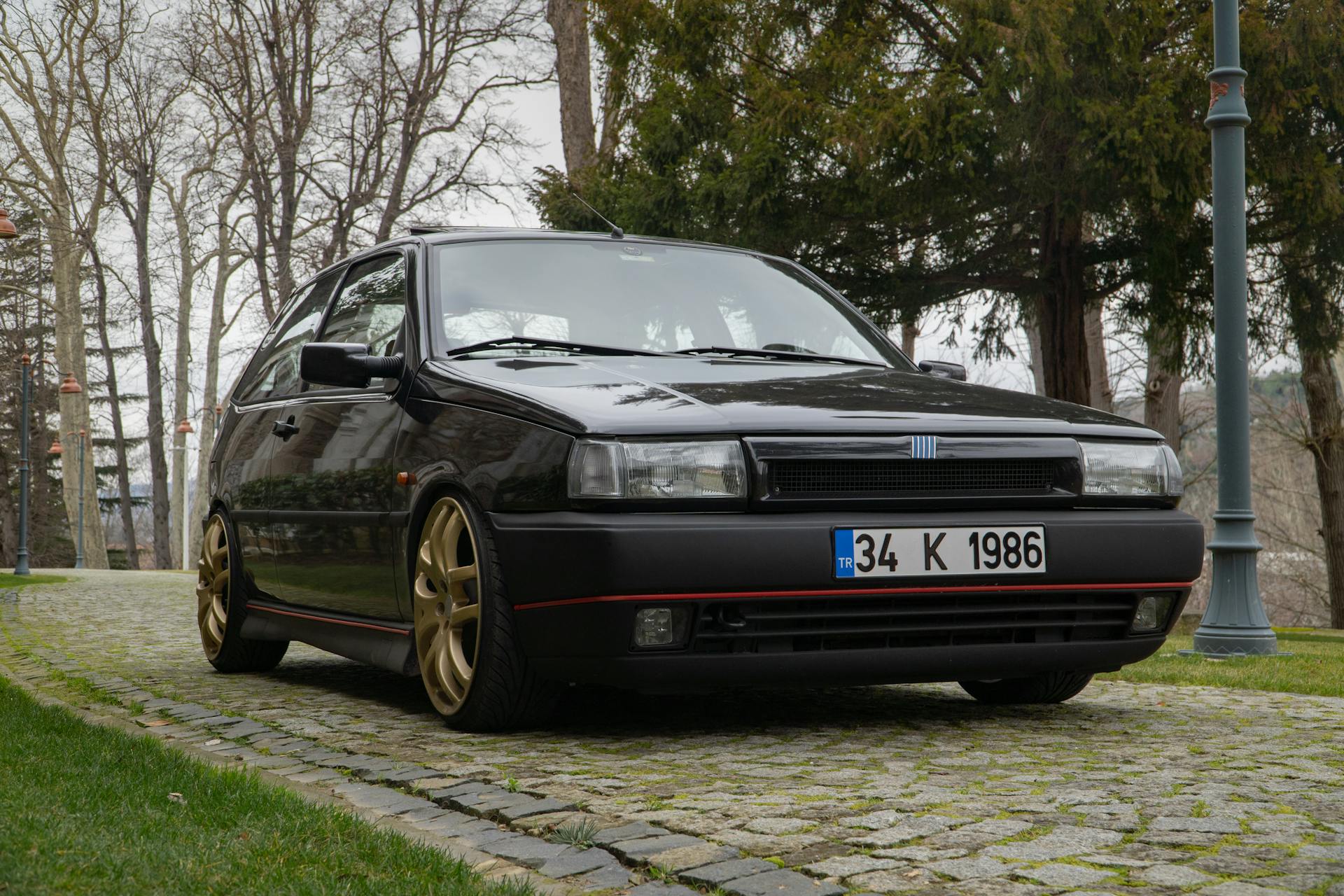 Classic black car with gold wheels on cobblestone, surrounded by trees.