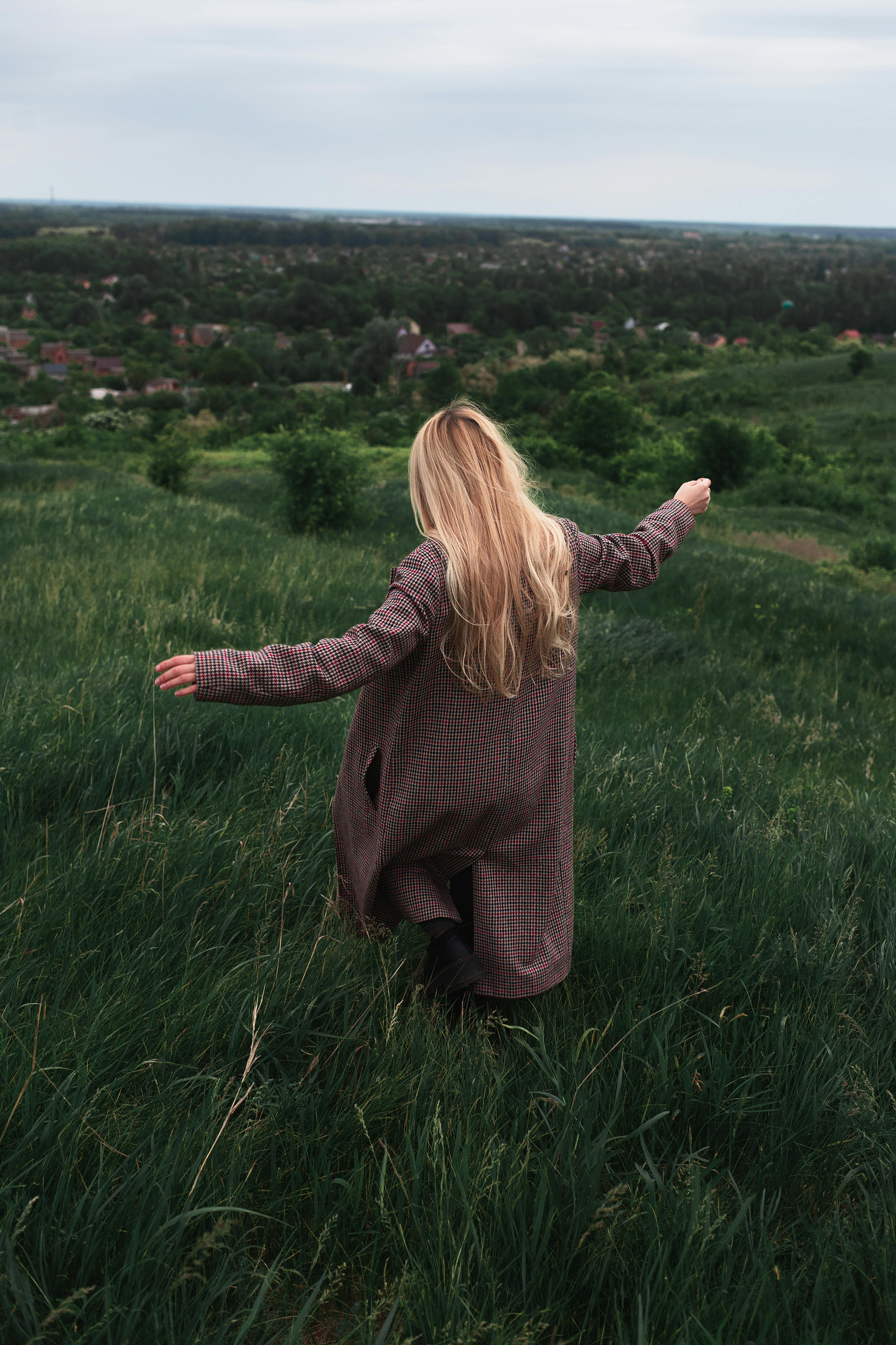 A blonde woman in a coat walks through a lush green field overlooking a village below.