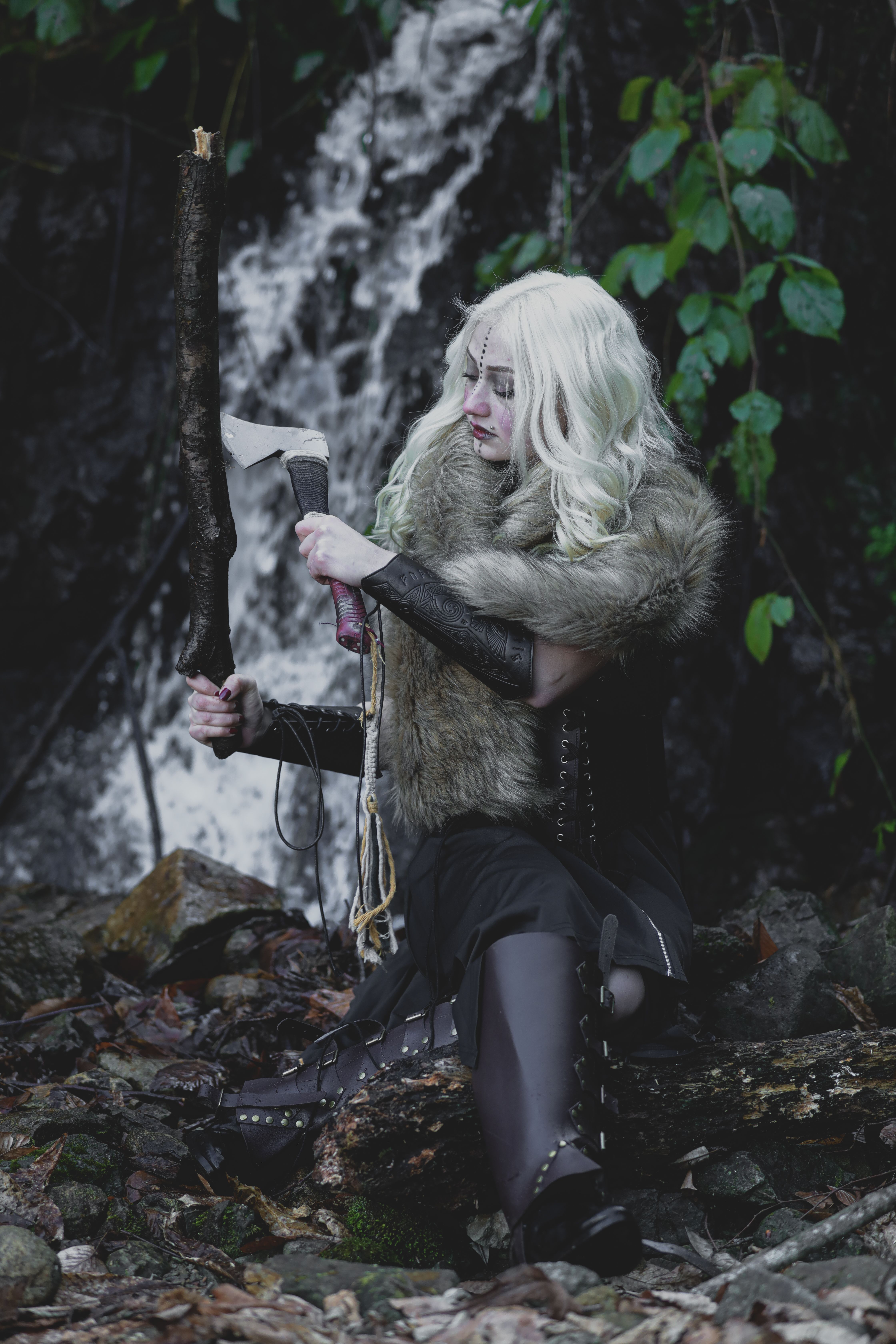 Blonde warrior sitting by waterfall in forest, crafting with tools.
