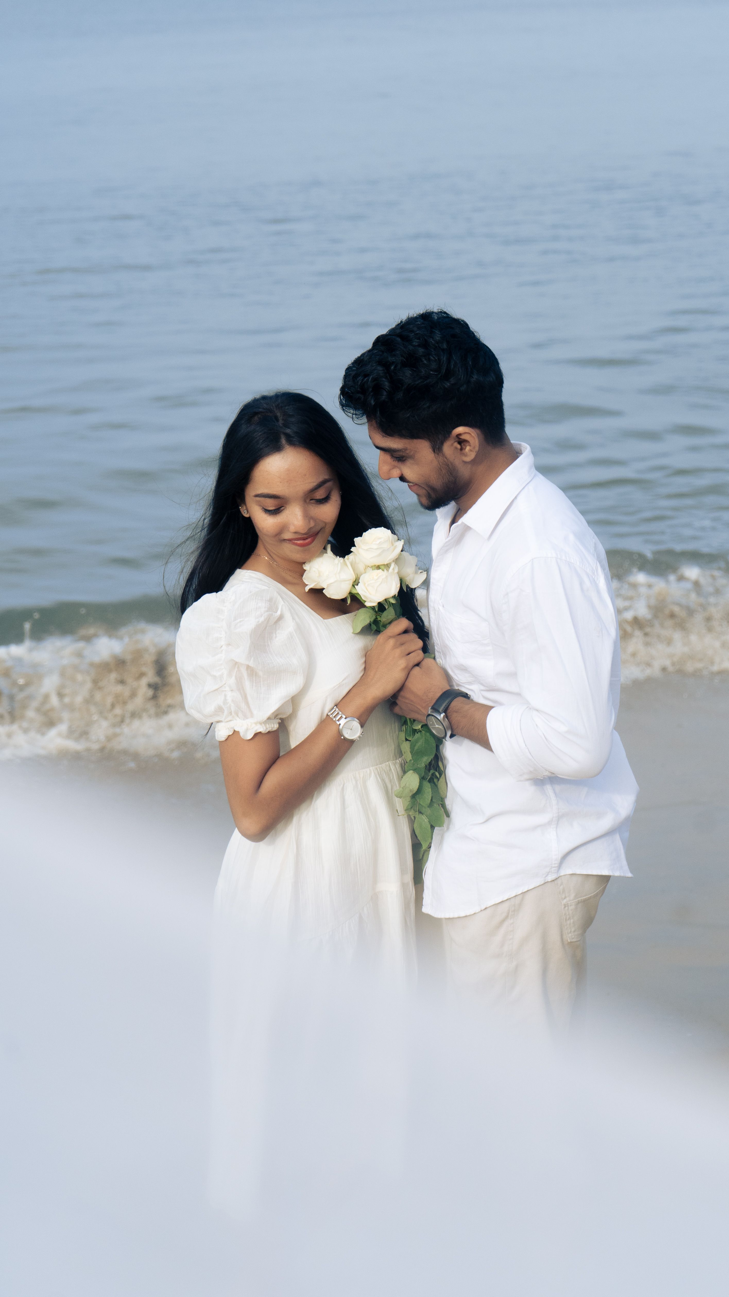 A loving couple shares a tender moment with white roses by the seaside.