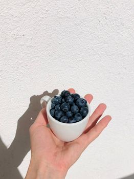 A hand holding a ceramic cup filled with fresh blueberries against a white textured wall.