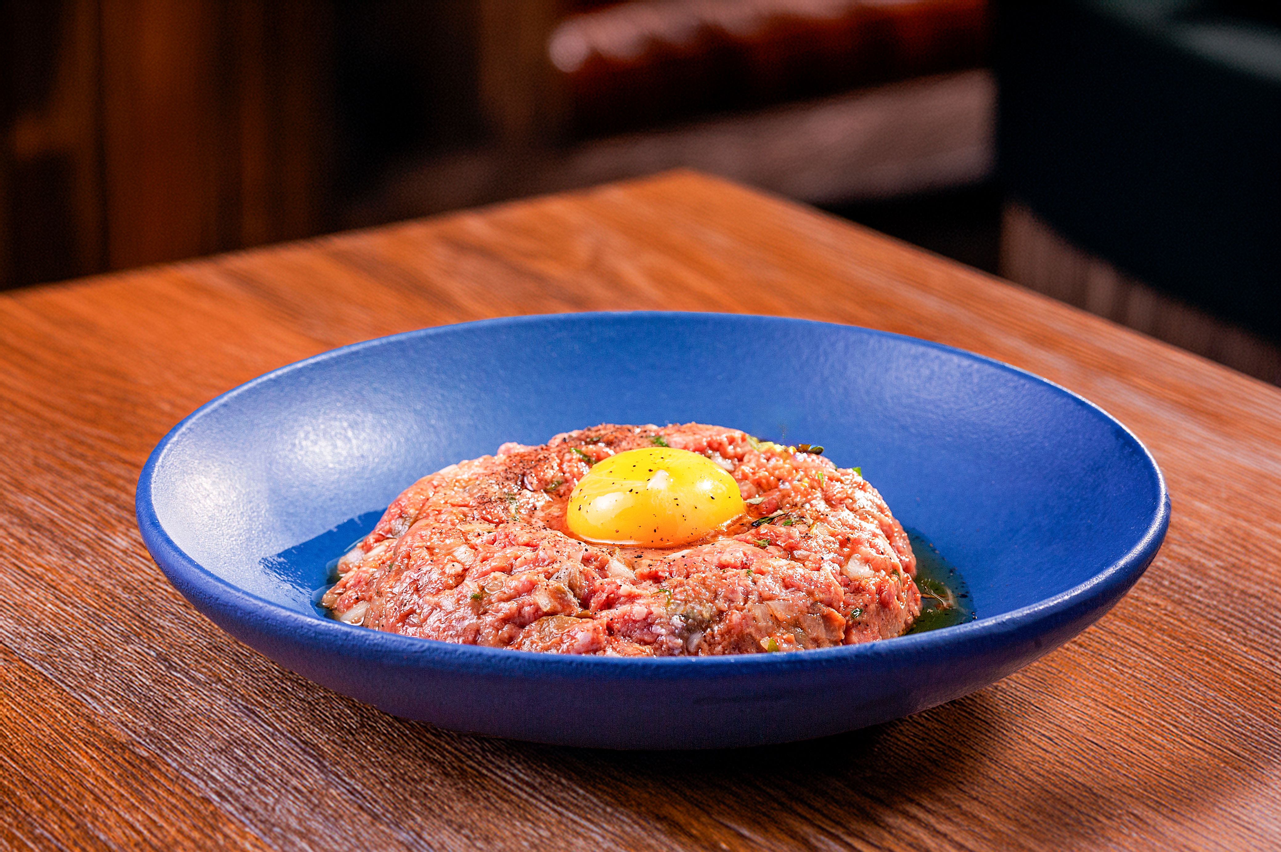 Appetizing beef tartare topped with an egg yolk served in a modern blue dish on a wooden table.
