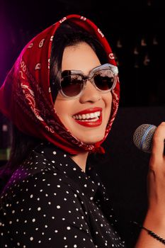 Smiling woman with sunglasses and headscarf holding a microphone at a music event.