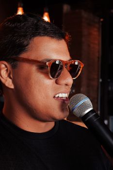 Young man in sunglasses singing passionately into a microphone indoors with ambient lighting.