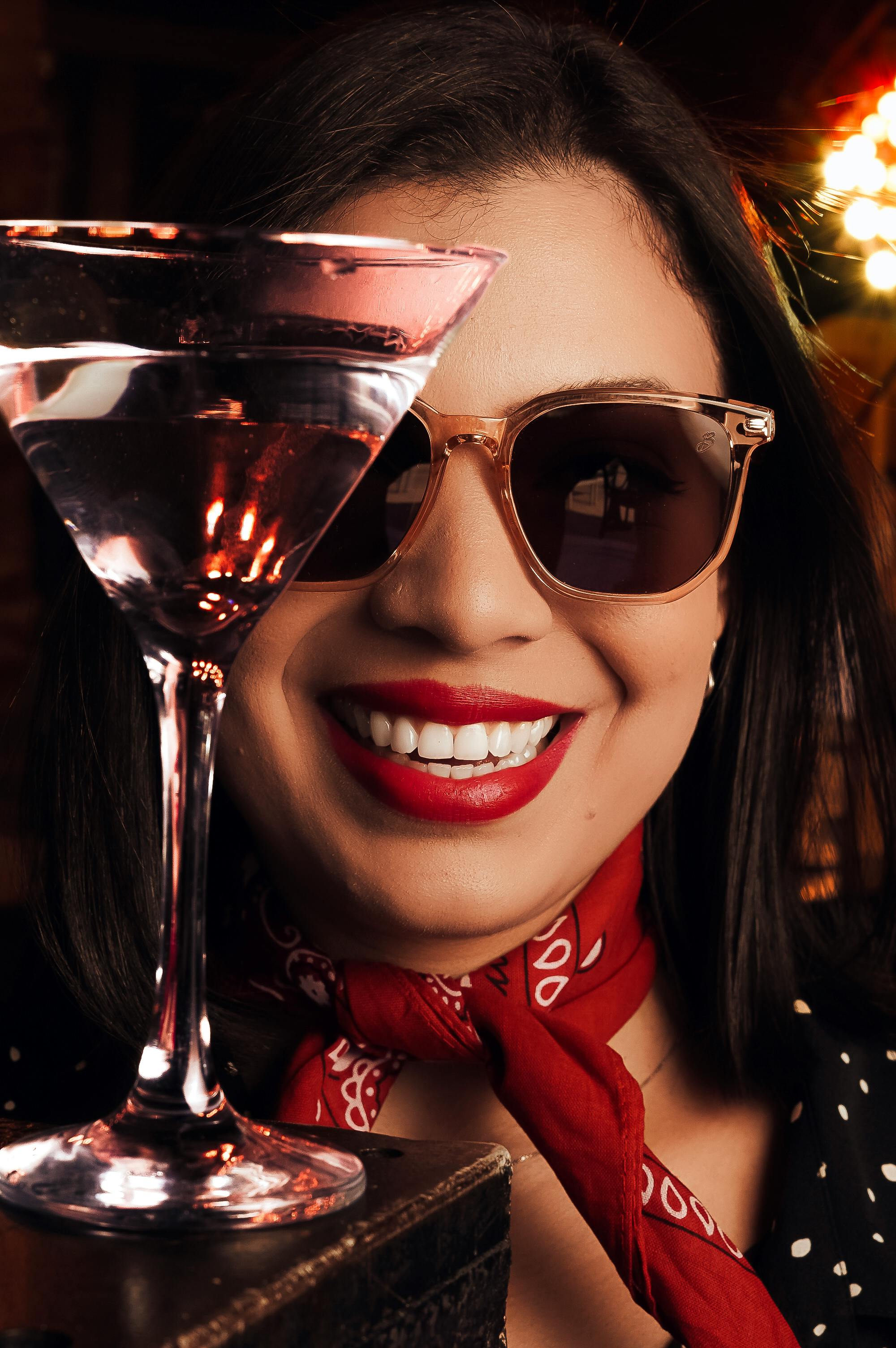 A fashionable woman with sunglasses and a red scarf enjoying a martini at a bar setting.