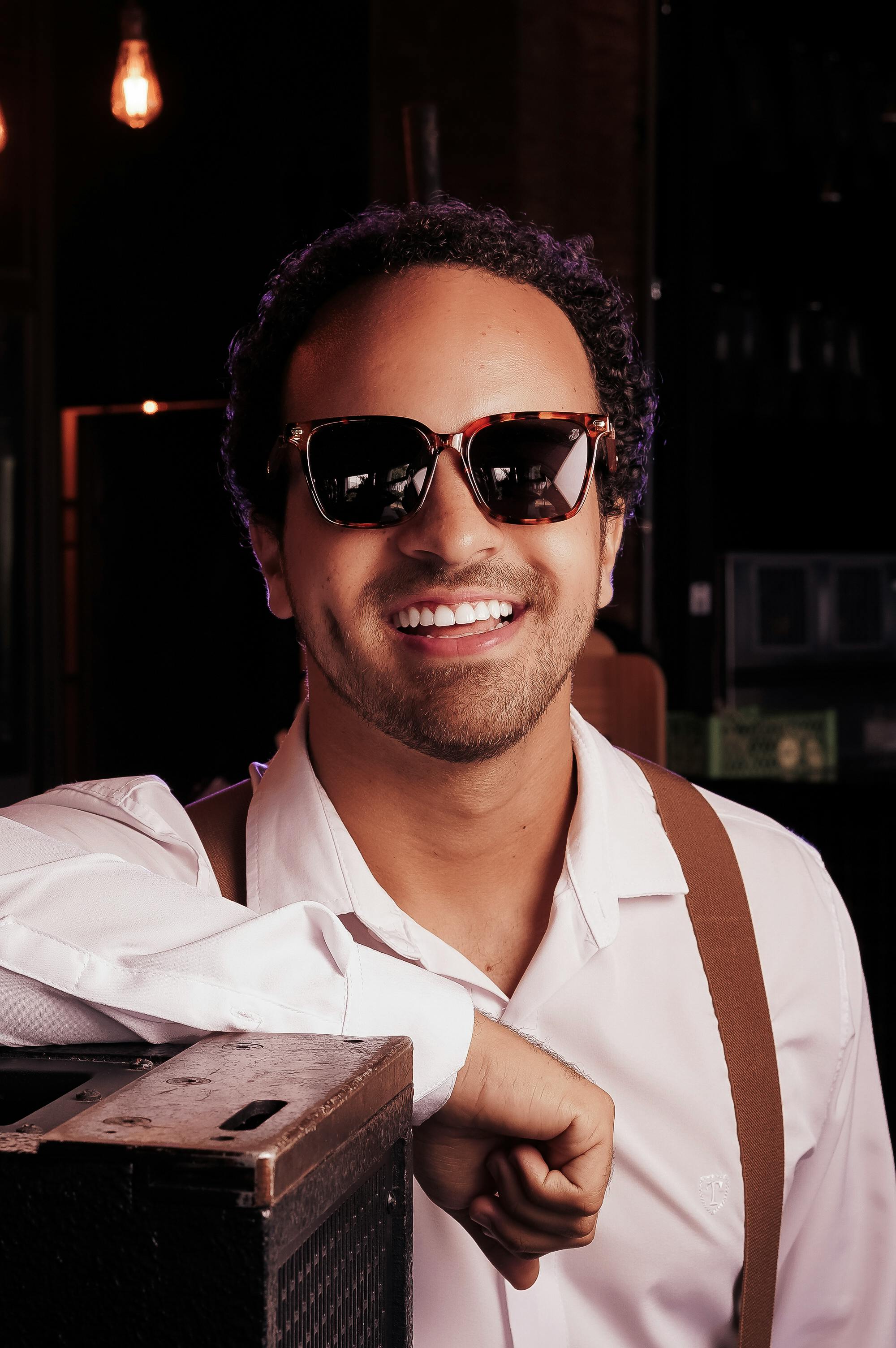 A cheerful man in suspenders and sunglasses, smiling indoors at a bar.