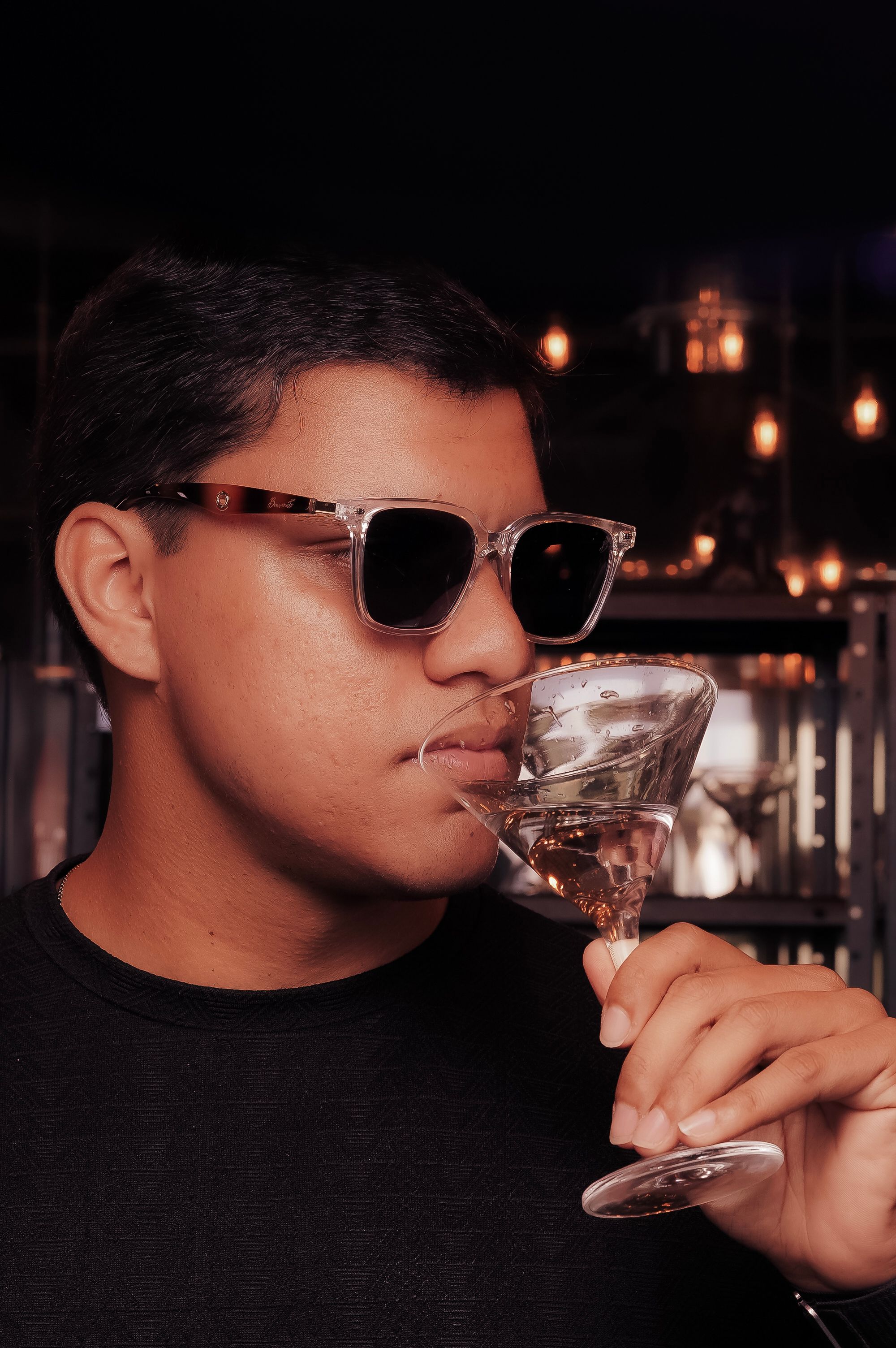 Young man wearing sunglasses enjoying a cocktail in a stylish bar setting.