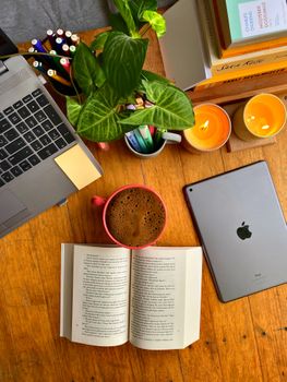Warm workspace with coffee, laptop, tablet, and books on a wooden table.