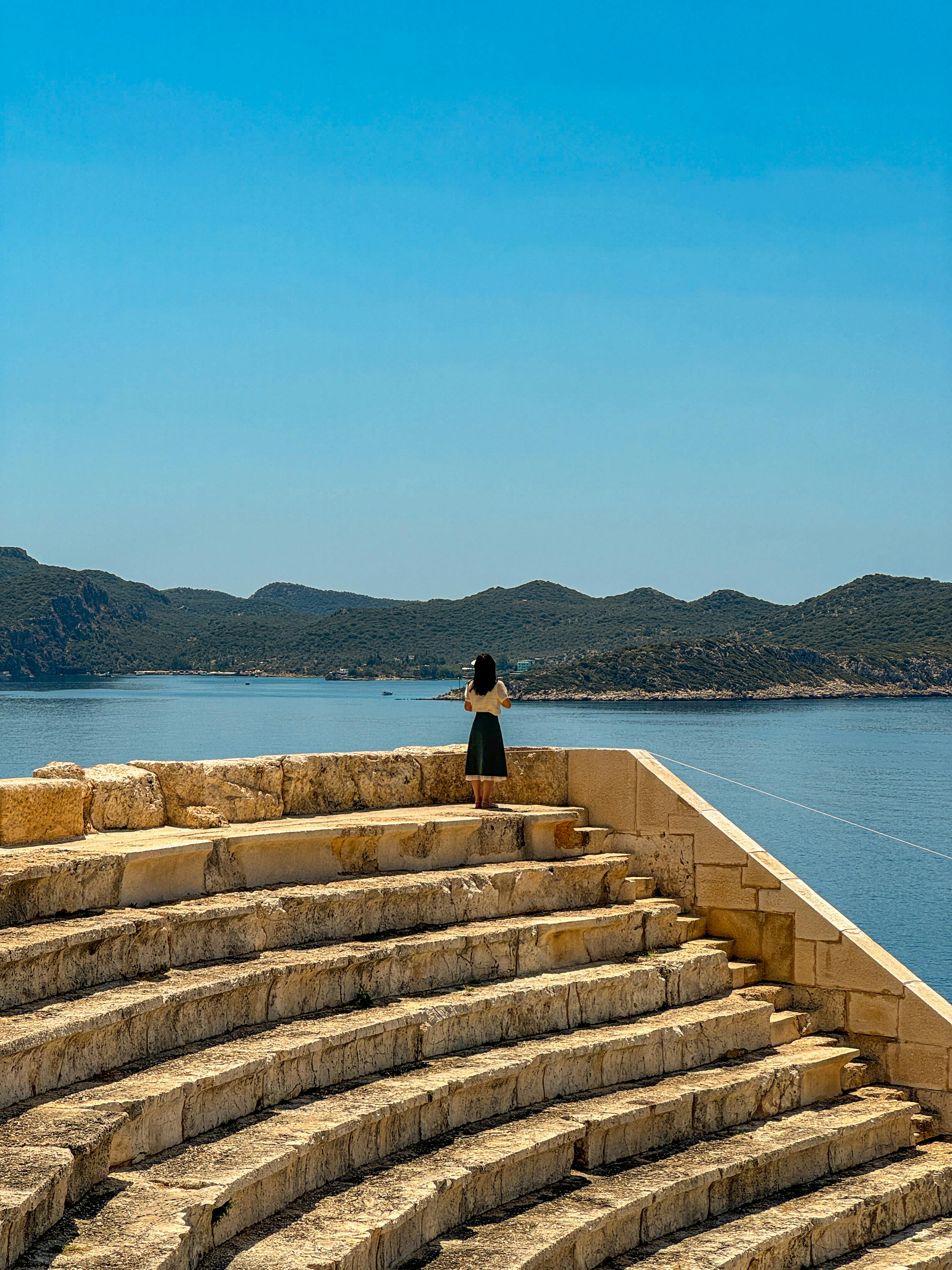 Free A woman standing on ancient steps, gazing at a beautiful coastal view under a clear blue sky. Stock Photo
