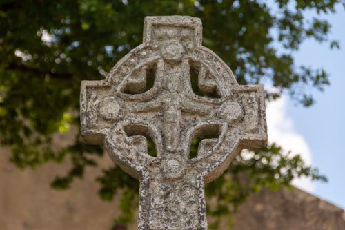 Free Close-up of an ancient Celtic cross against a lush green tree and blue sky background. Stock Photo