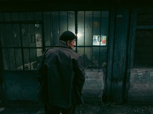 Free An elderly man peers into a dimly lit shop through a window at night, creating a mysterious urban scene. Stock Photo