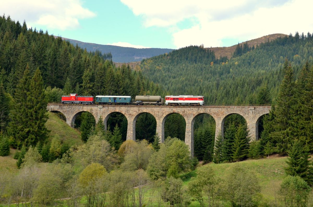 Două locomotive roșii traversând viaductul de piatră Chmarošský din Slovacia, între brazi