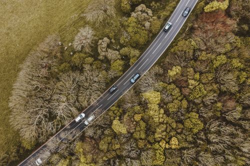 Free Drone view captures vehicles driving on a winding road through a lush forest, showcasing the blend of nature and transportation. Stock Photo