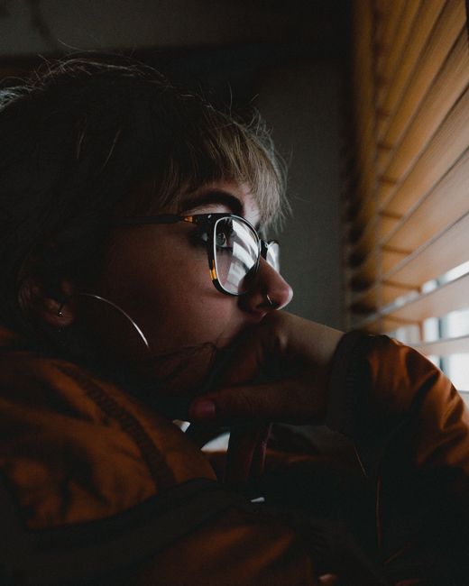Pensive woman in eyeglasses gazing through blinds, contemplating indoors.