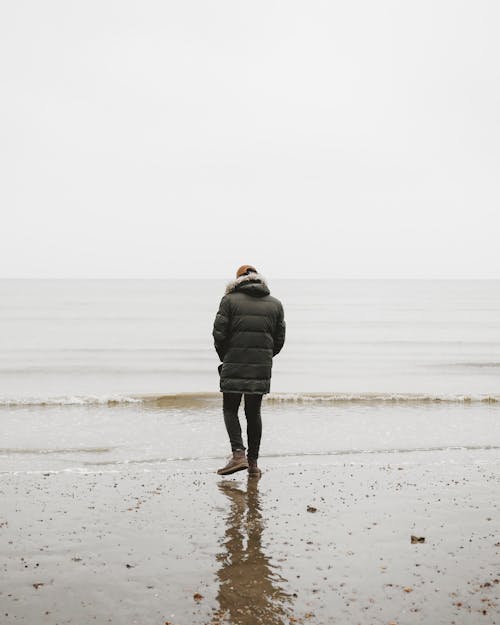 Free Back View of a Person Wearing Black Bubble Jacket Walking Towards the Sea Stock Photo