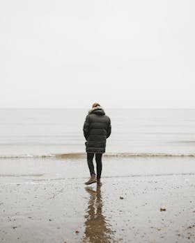 一个穿着泡泡夹克的人独自走在宁静的海滩上，面对平静的大海，透着孤独