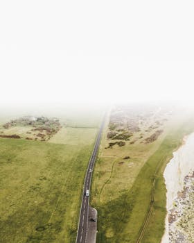 无人机从高空俯拍，一条风景优美的沿海道路蜿蜒伸展，道路两侧是郁郁葱葱的绿色植物和引人注目的悬崖，映衬在多云的天空下