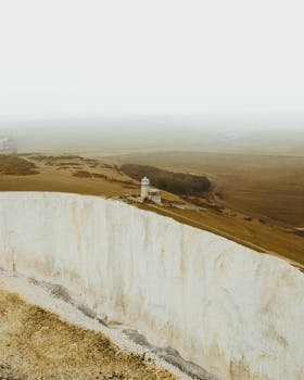 A foggy landscape featuring a lighthouse on dramatic cliffs by the sea.