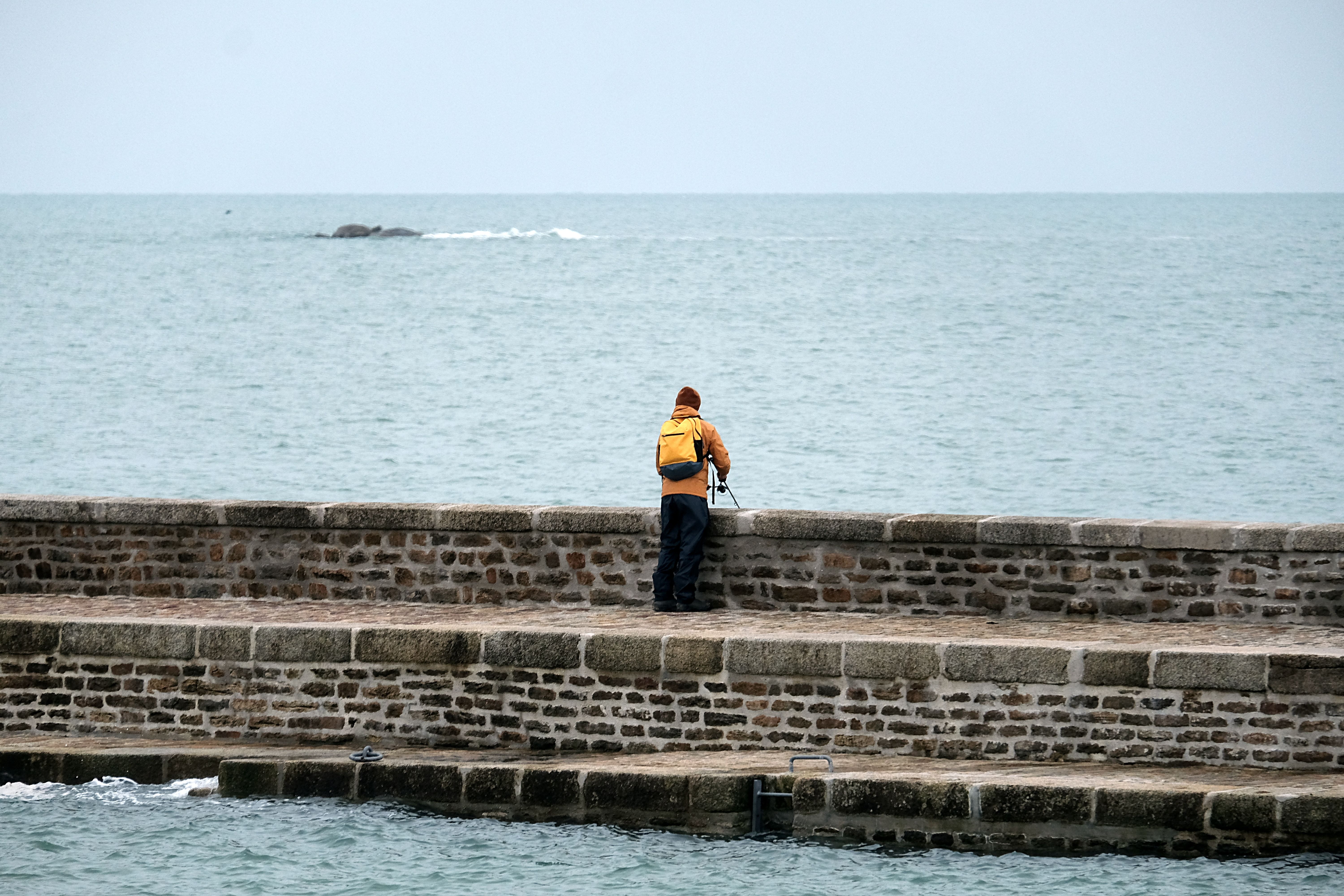 A lone fisherman in vibrant clothes stands on a stone pier against the vast sea.