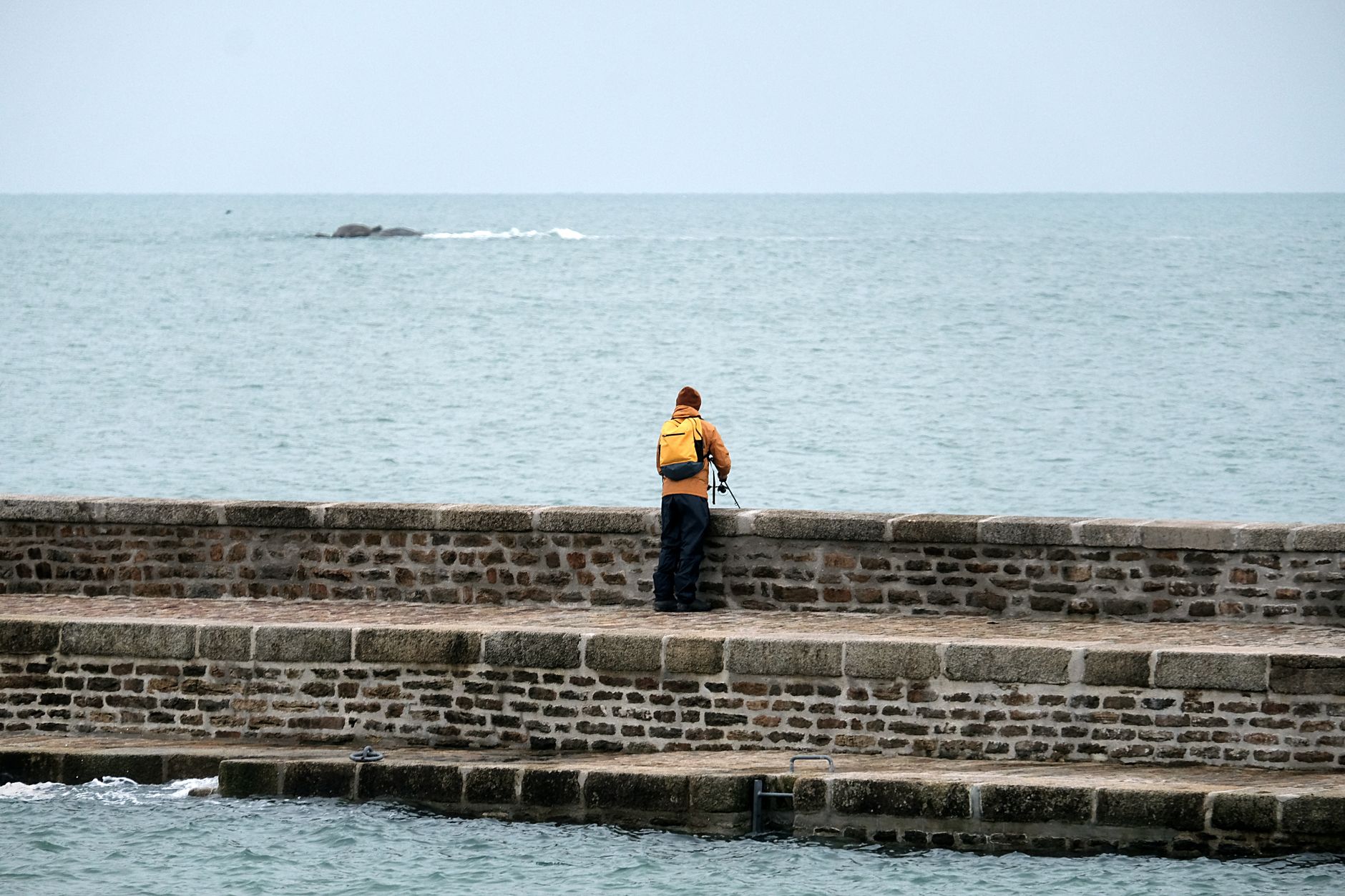 A lone fisherman in vibrant clothes stands on a stone pier against the vast sea.