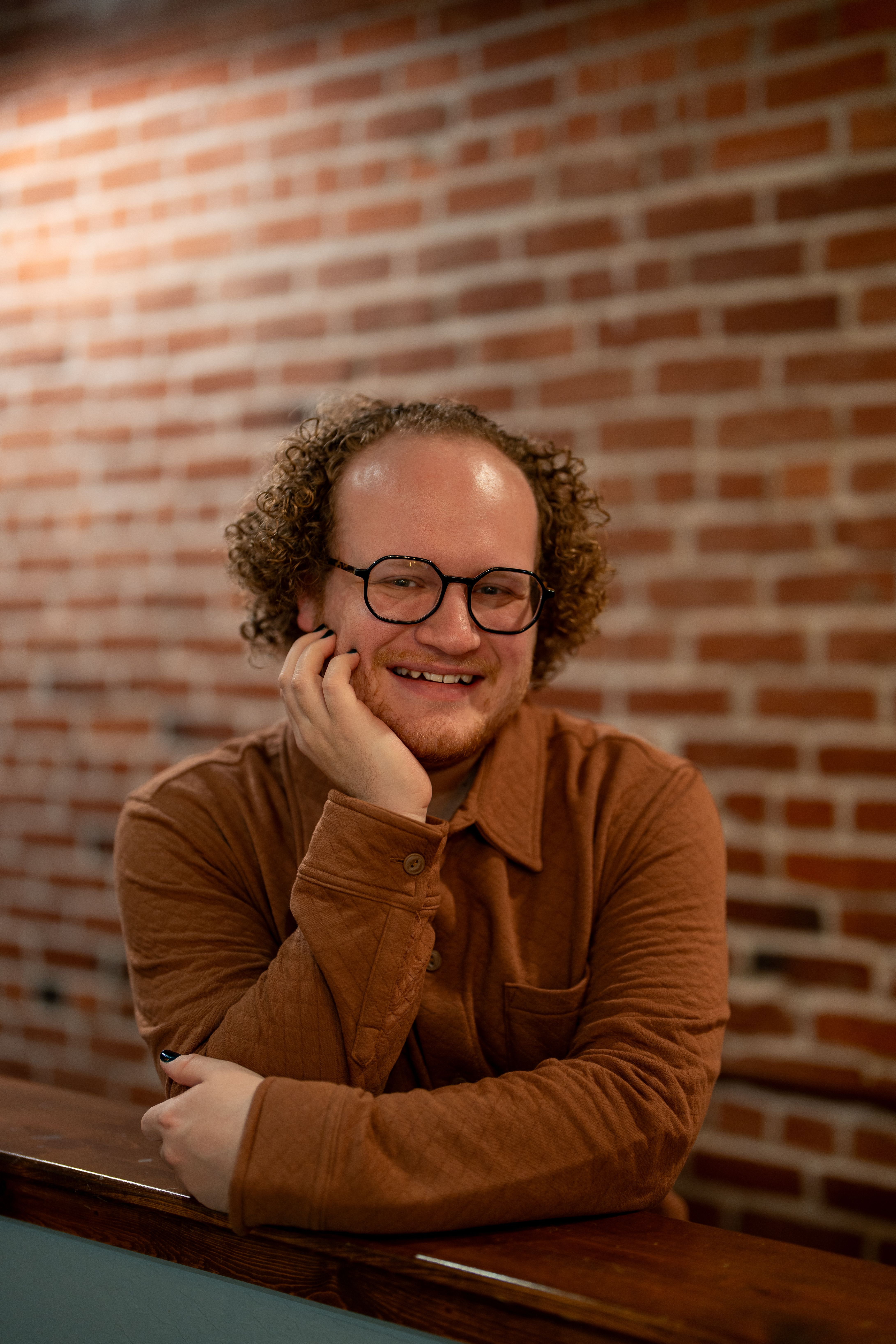 A cheerful man with curly hair and glasses smiles warmly indoors against a brick wall.