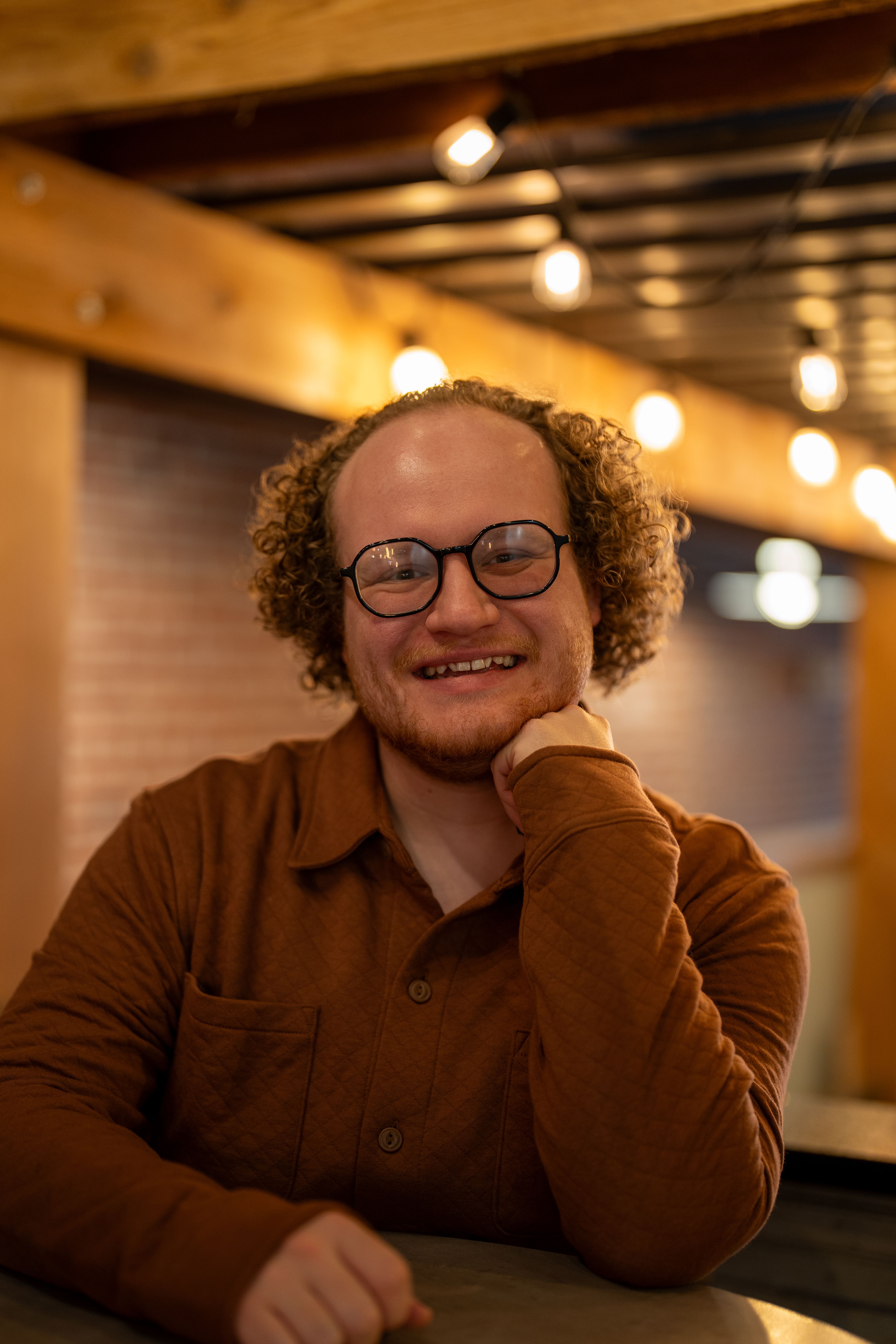 Smiling man with curly hair in a cozy setting, wearing glasses and a brown shirt.