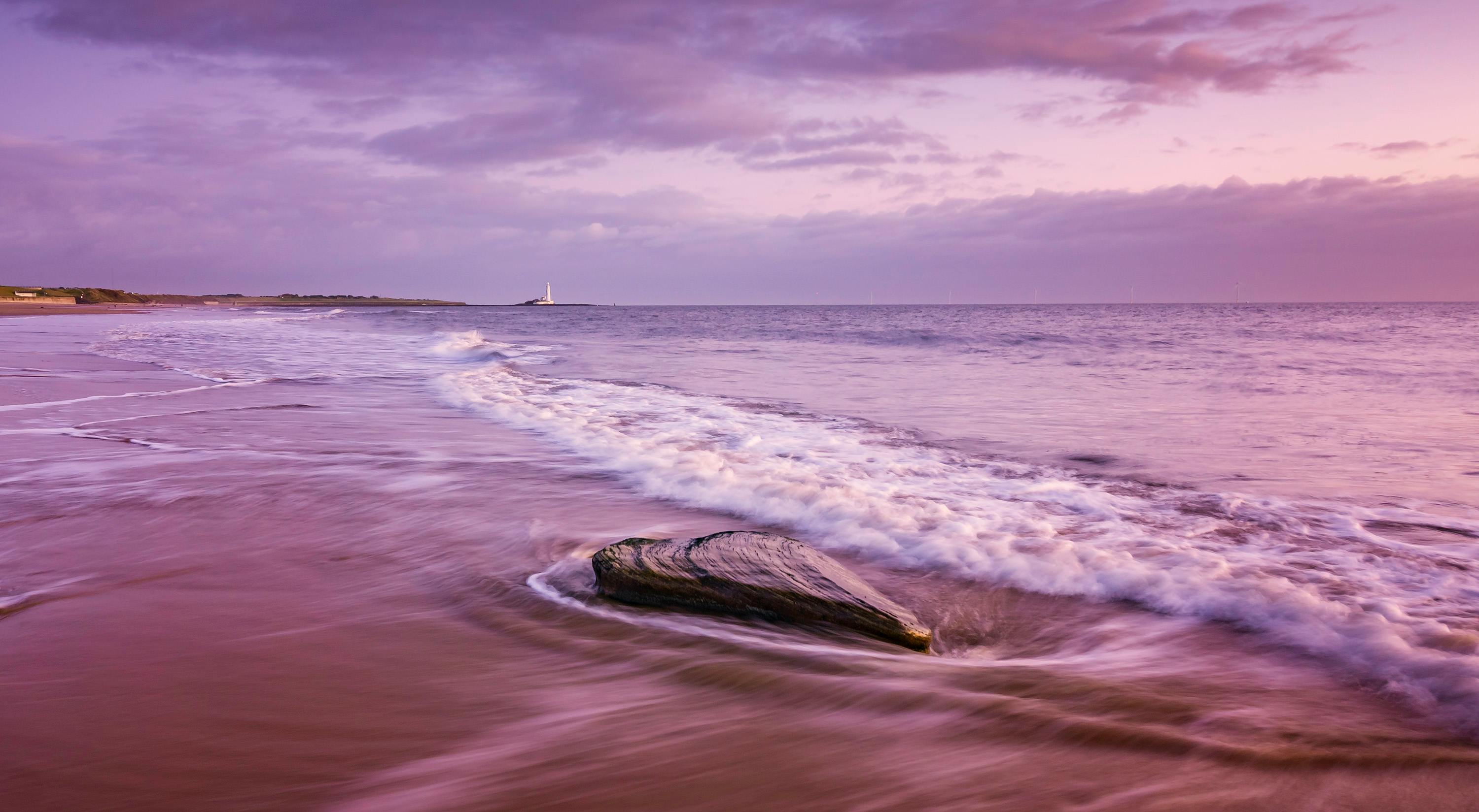日出时分，宁静的海滩景色中，海浪与远处的灯塔共同捕捉了这份静谧之美
