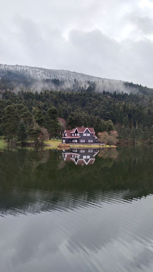 Free A serene house reflecting on a calm lake with a backdrop of foggy forest and mountains. Stock Photo