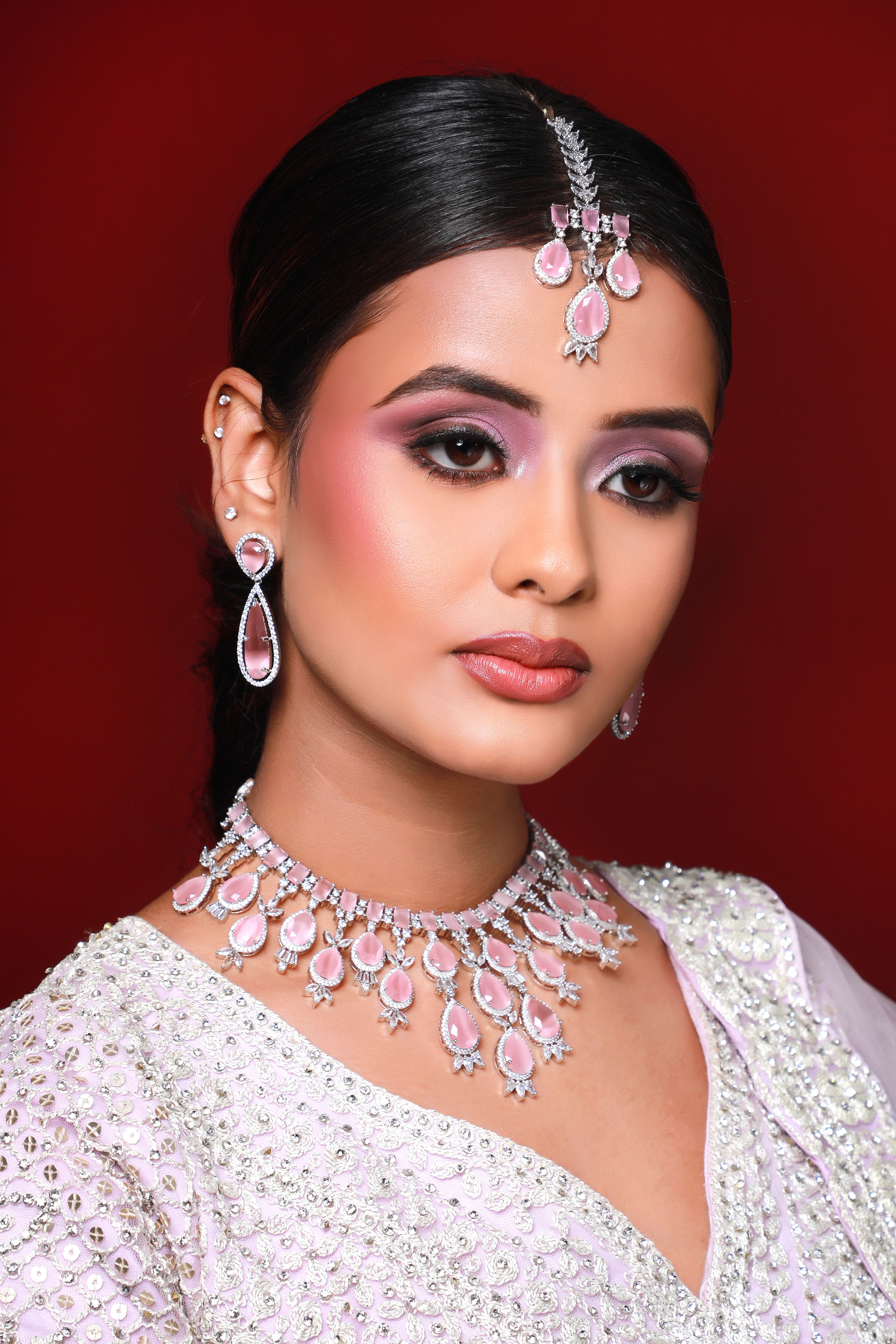 Close-up portrait of a young woman adorned in South Asian bridal jewelry and makeup on a red background.