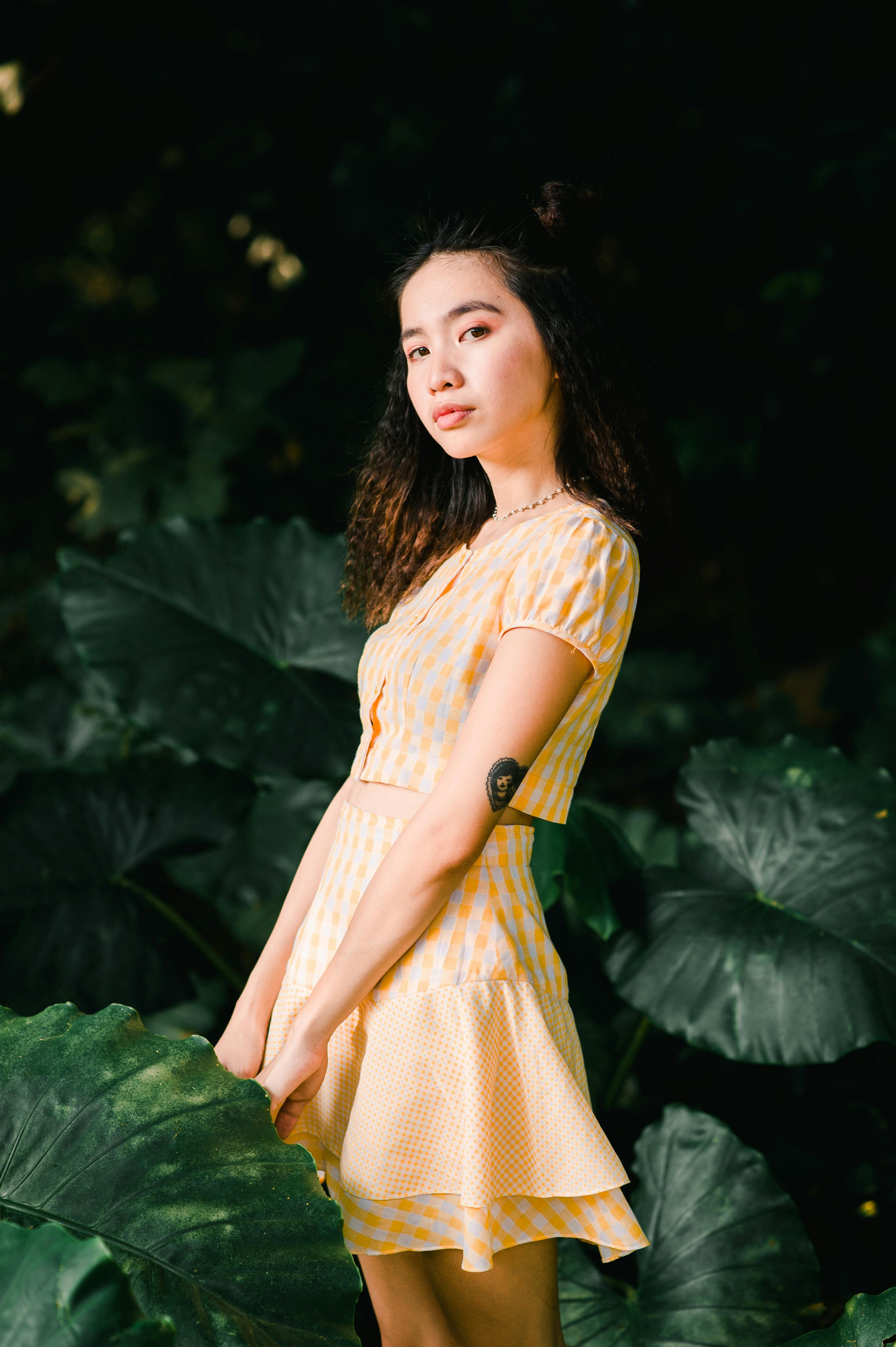 A stylish woman in a yellow dress posing against a backdrop of lush green leaves outdoors.