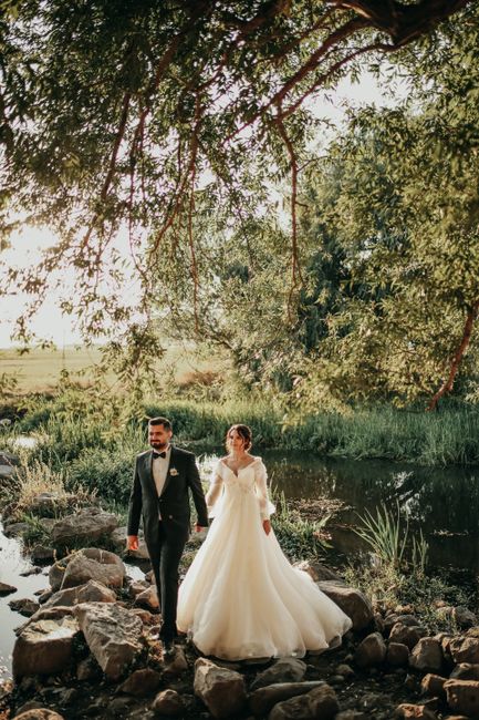 A beautiful couple in wedding attire standing by a serene river under lush green trees.