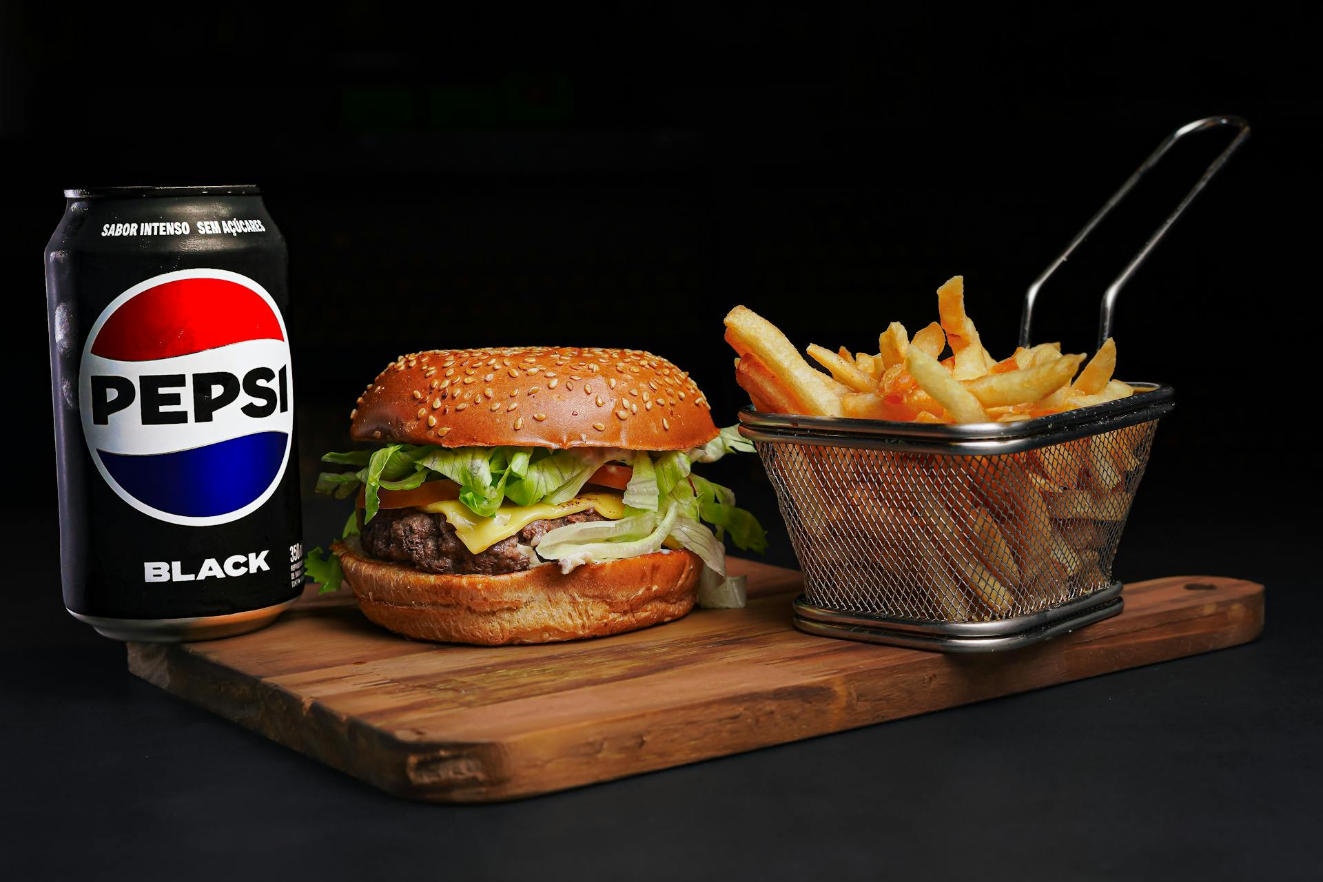 Close-up of a burger with fries and a Pepsi Black can on a wooden board.