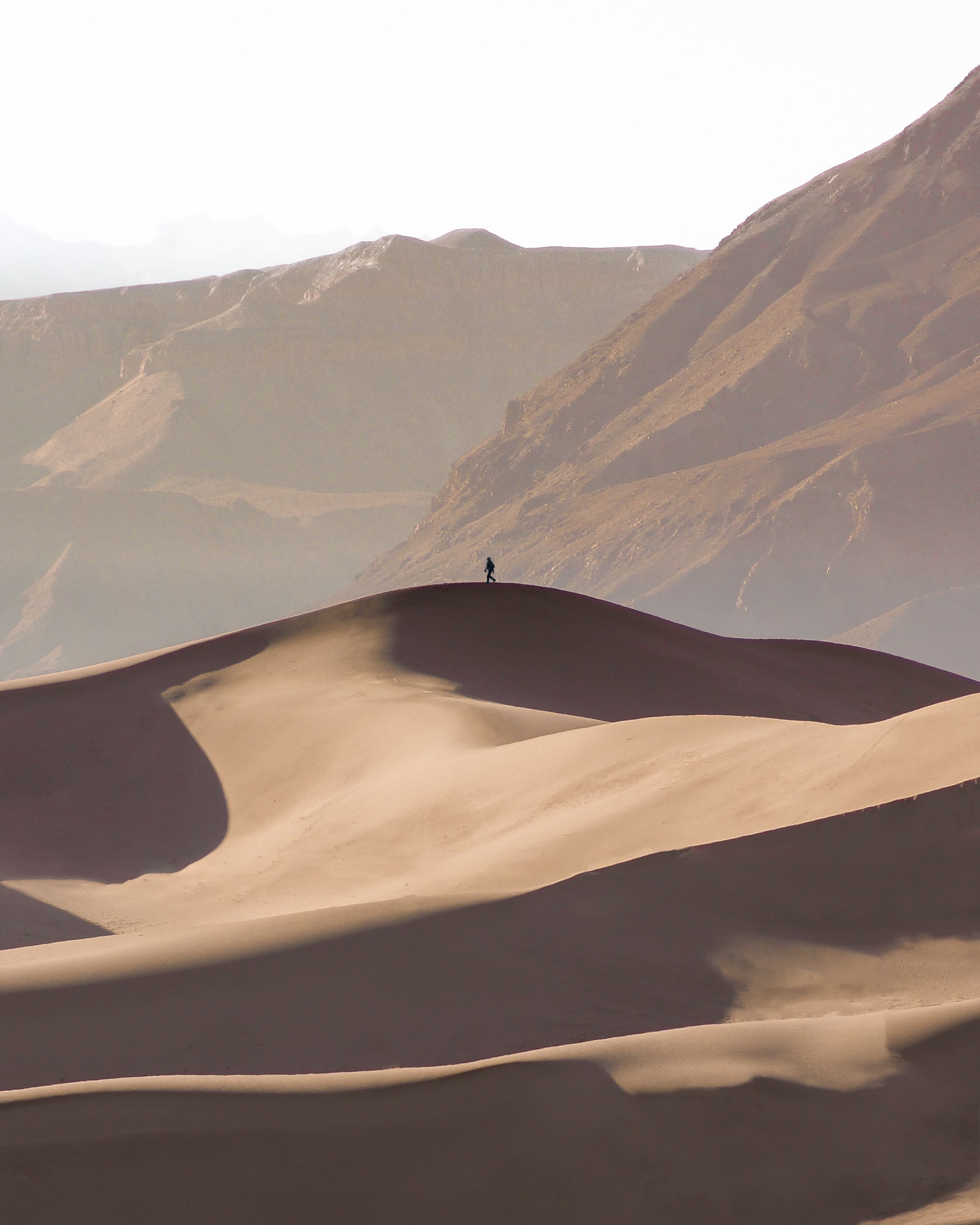 A lone person stands on expansive sand dunes beneath towering mountains in a vast desert.