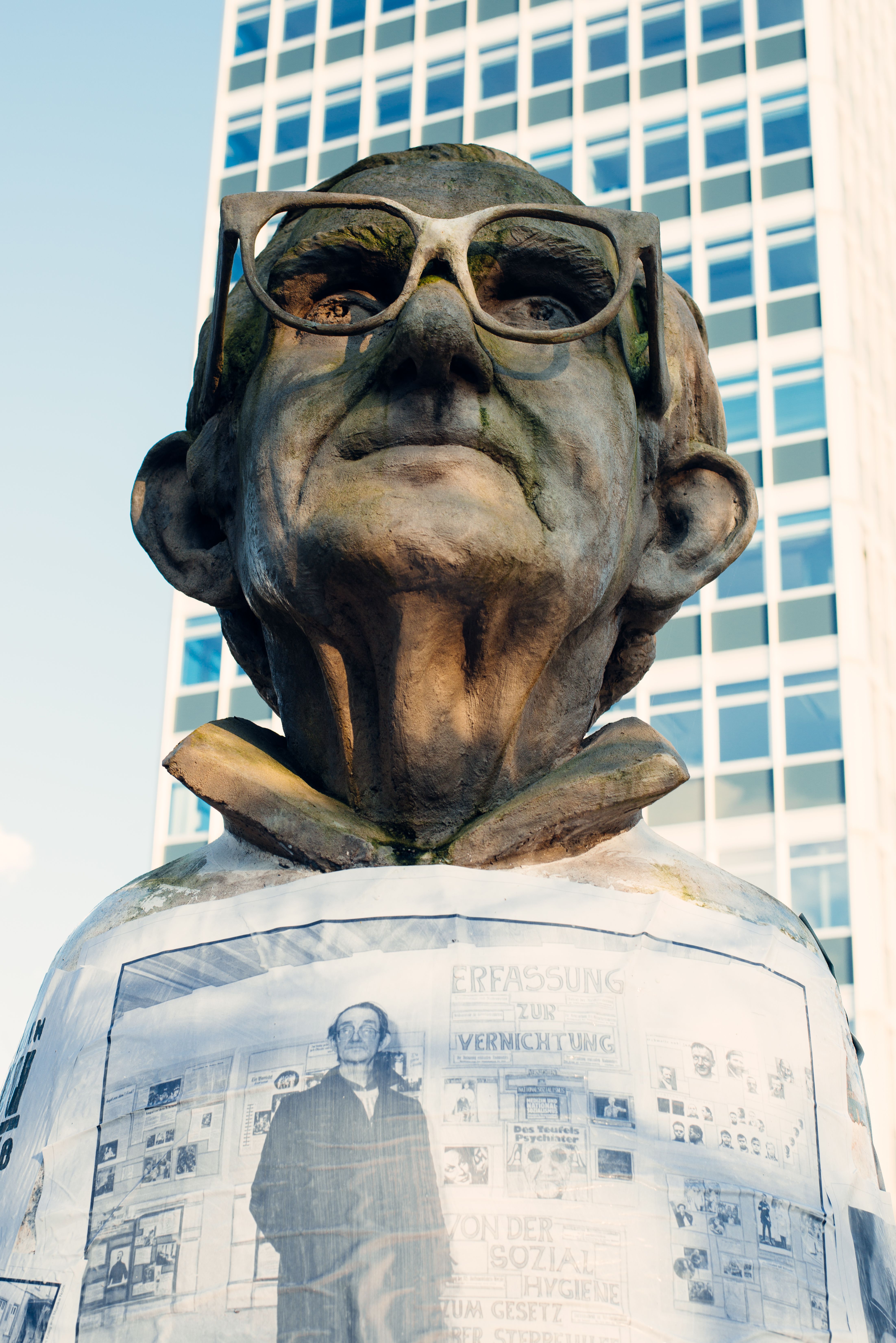Sculpture of a person with glasses in front of a high-rise building, daytime.