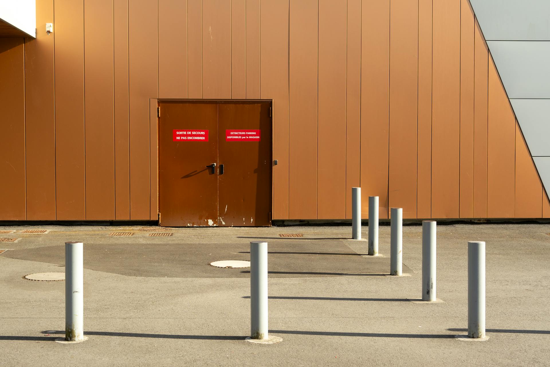 Exterior of an industrial building with brown emergency exit door, concrete bollards.