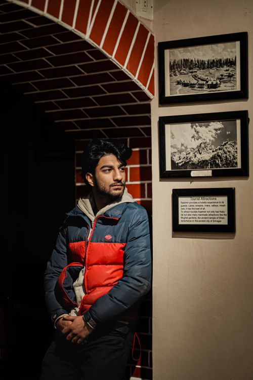 Free A young man in a colorful jacket leans against a wall with framed pictures indoors. Stock Photo