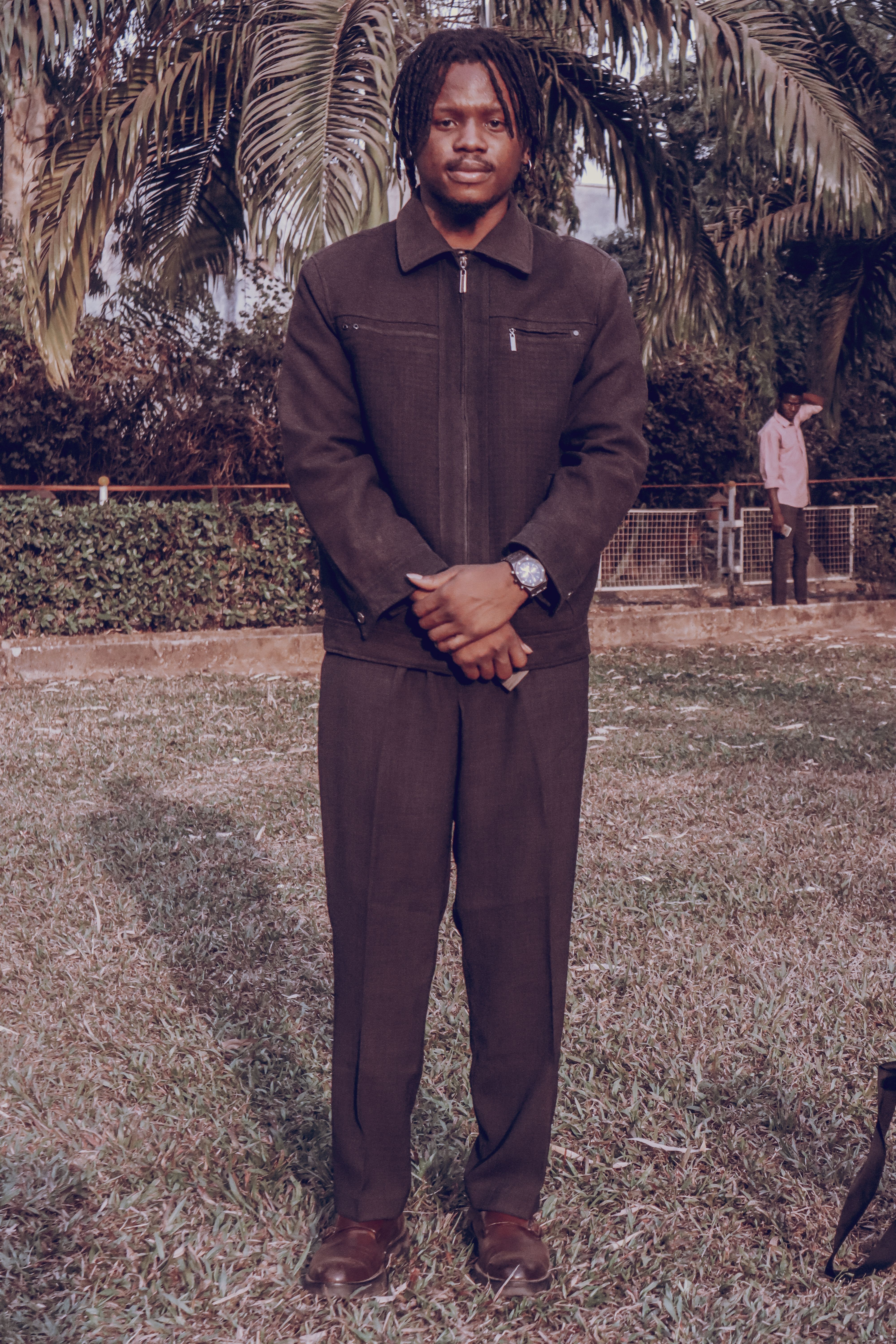 Stylish man in a dark suit standing on grass with tropical backdrop, daytime.