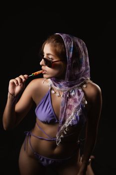Fashionable woman in bikini with scarf and sunglasses holding a lollipop.