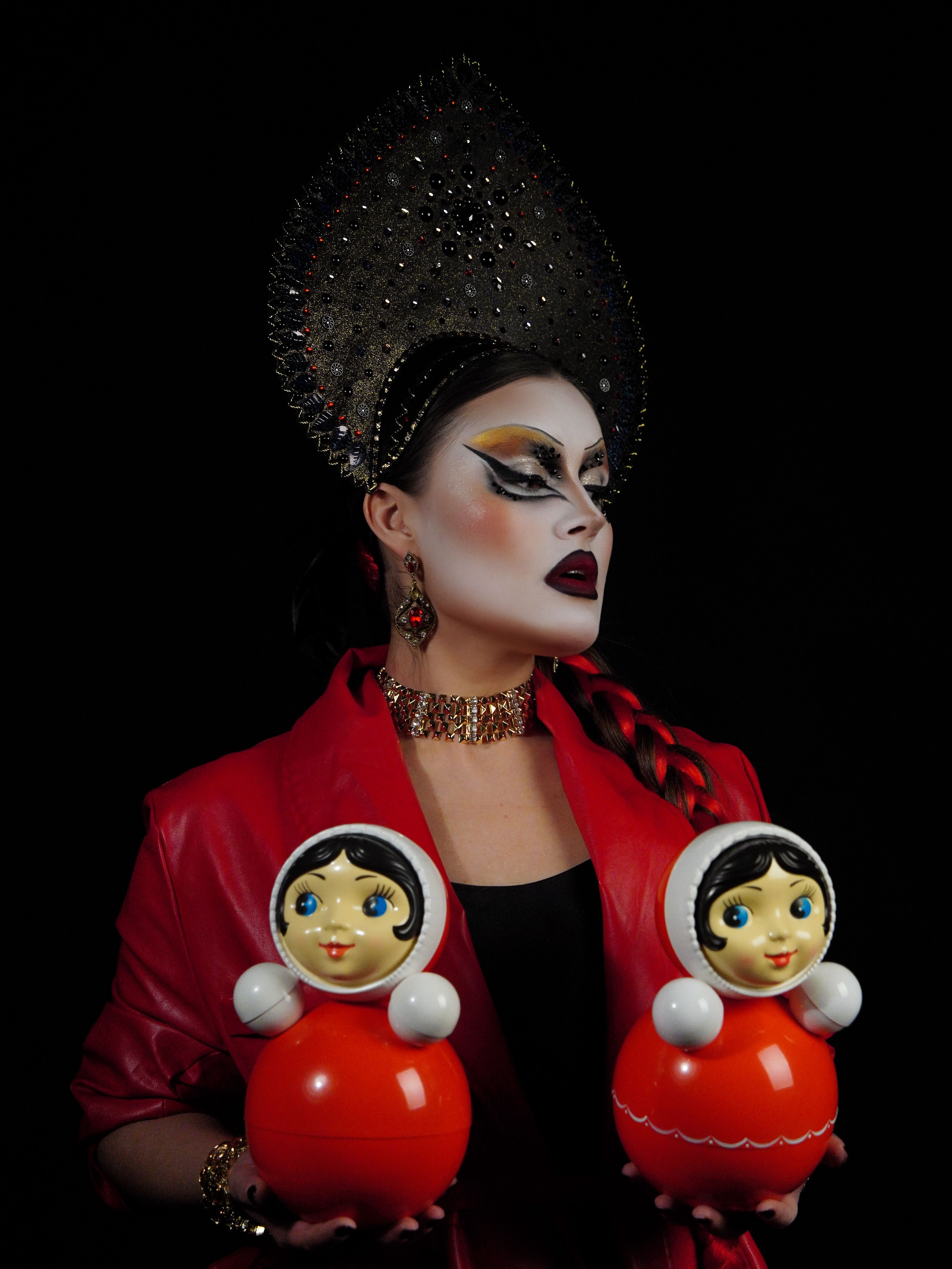 Drag queen in elaborate costume holding two traditional Russian dolls against a dark backdrop.