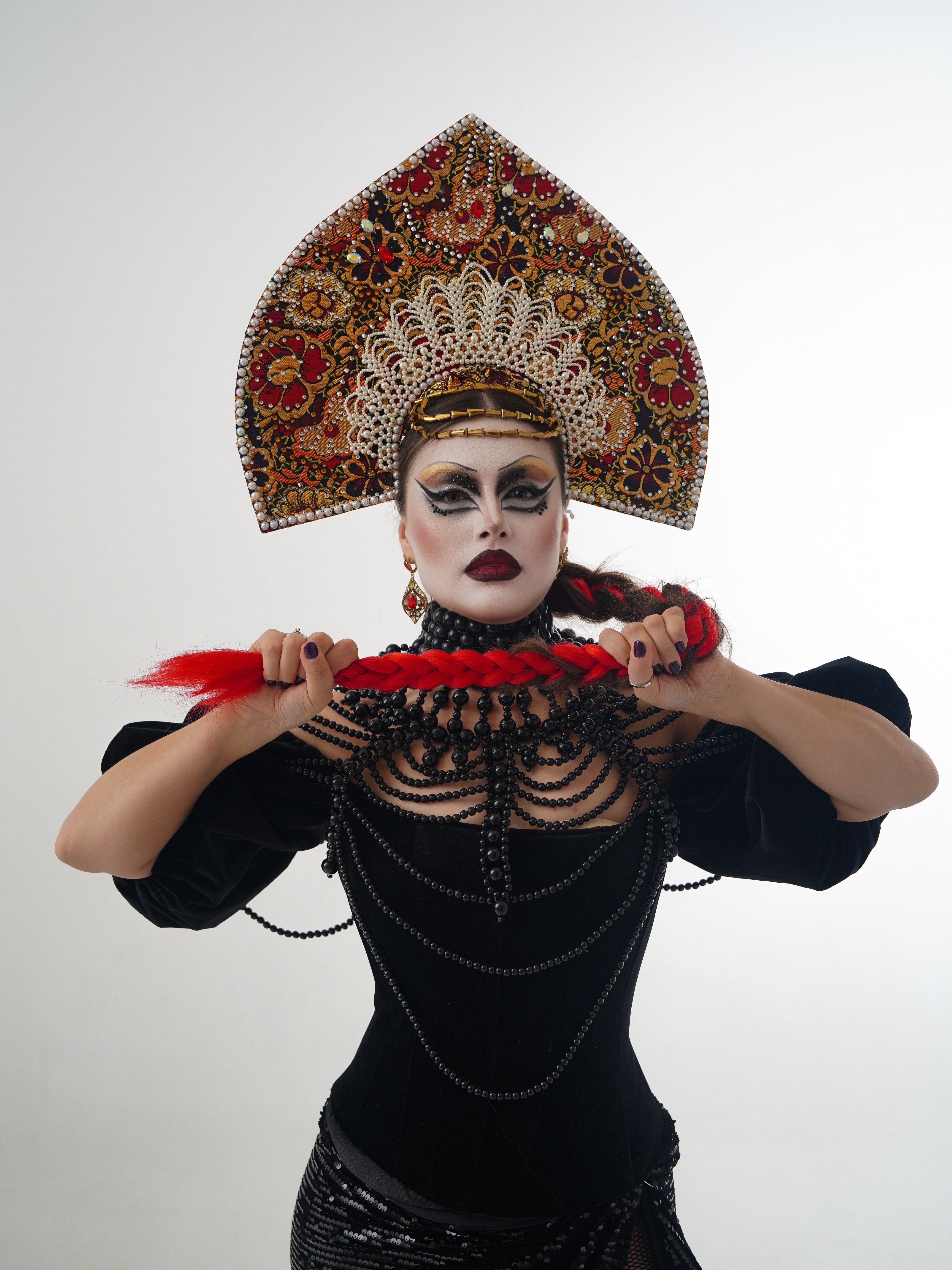 Portrait of a woman in ornate headdress and dramatic make-up holding a red braid.