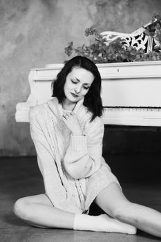 A woman in a cozy sweater poses playfully beside a white piano in a stylish black and white setting.