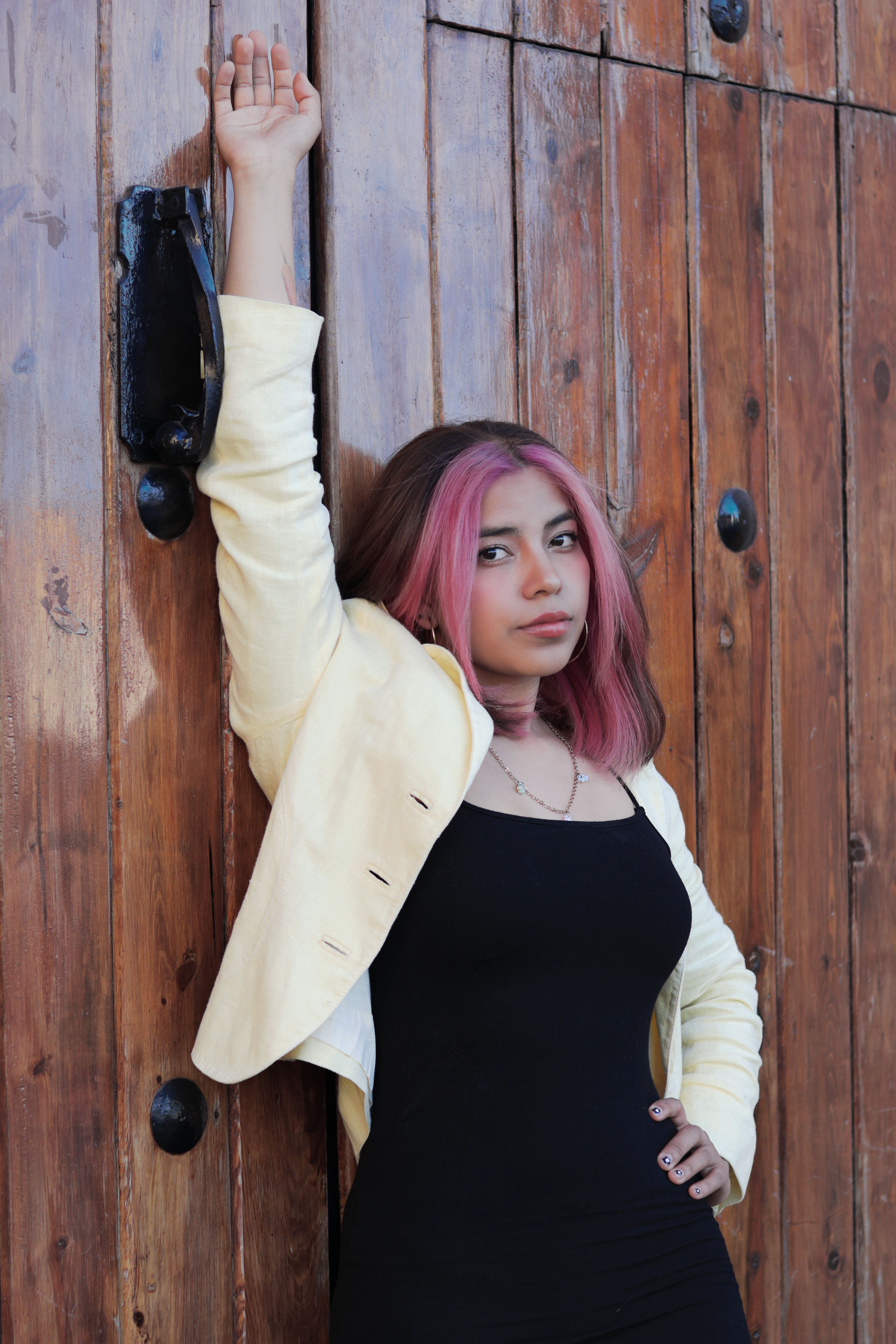A young woman with pink hair poses confidently against a rustic wooden door, wearing a black dress and white jacket.