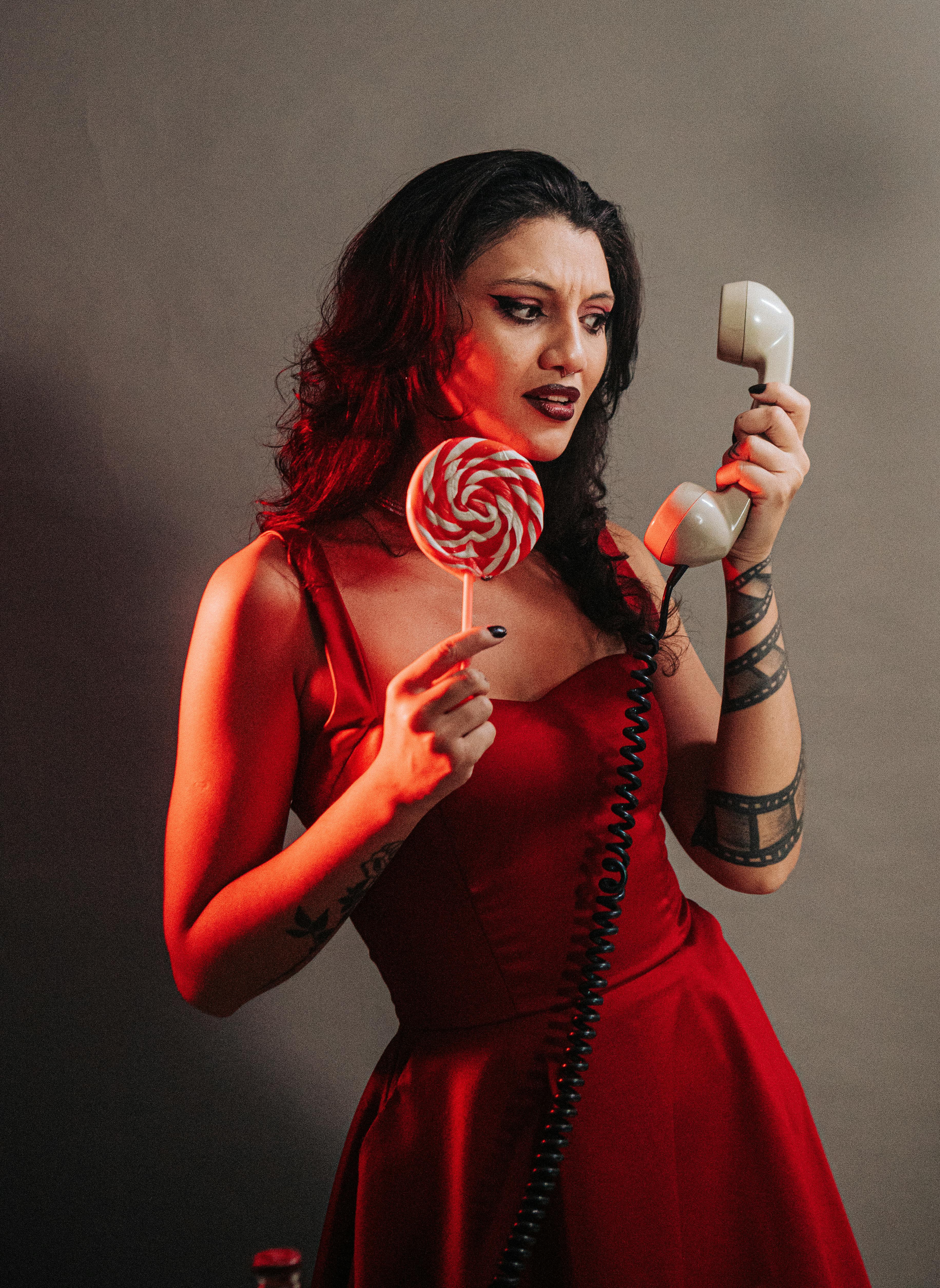 Miễn phí Bức chân dung ấn tượng của một người phụ nữ mặc váy đỏ, tay cầm một cây kẹo mút lớn và ống nghe điện thoại cổ điển. Ảnh lưu trữ