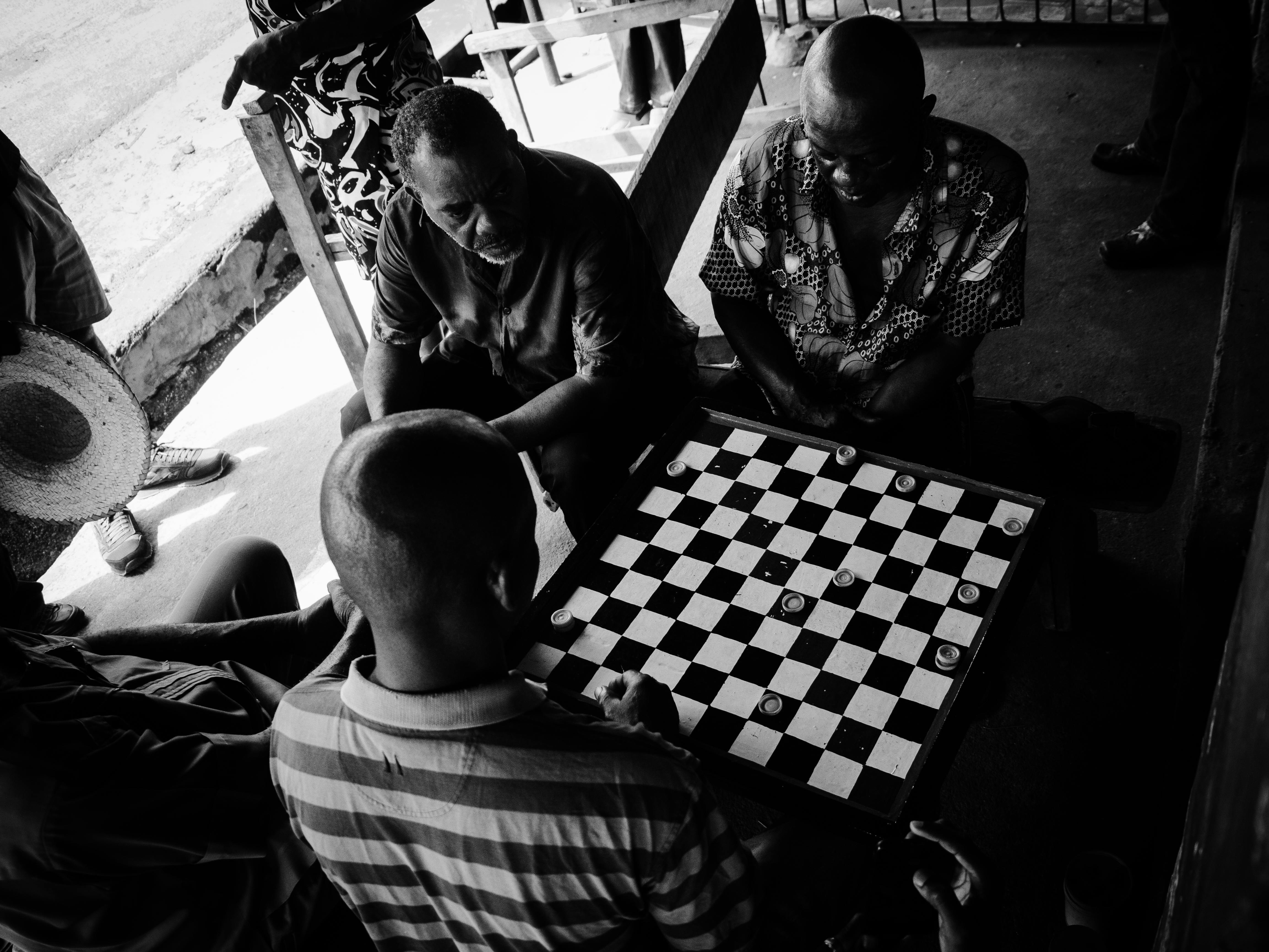 Men engaged in an outdoor checkers game in a lively, urban setting. Captured in black and white.