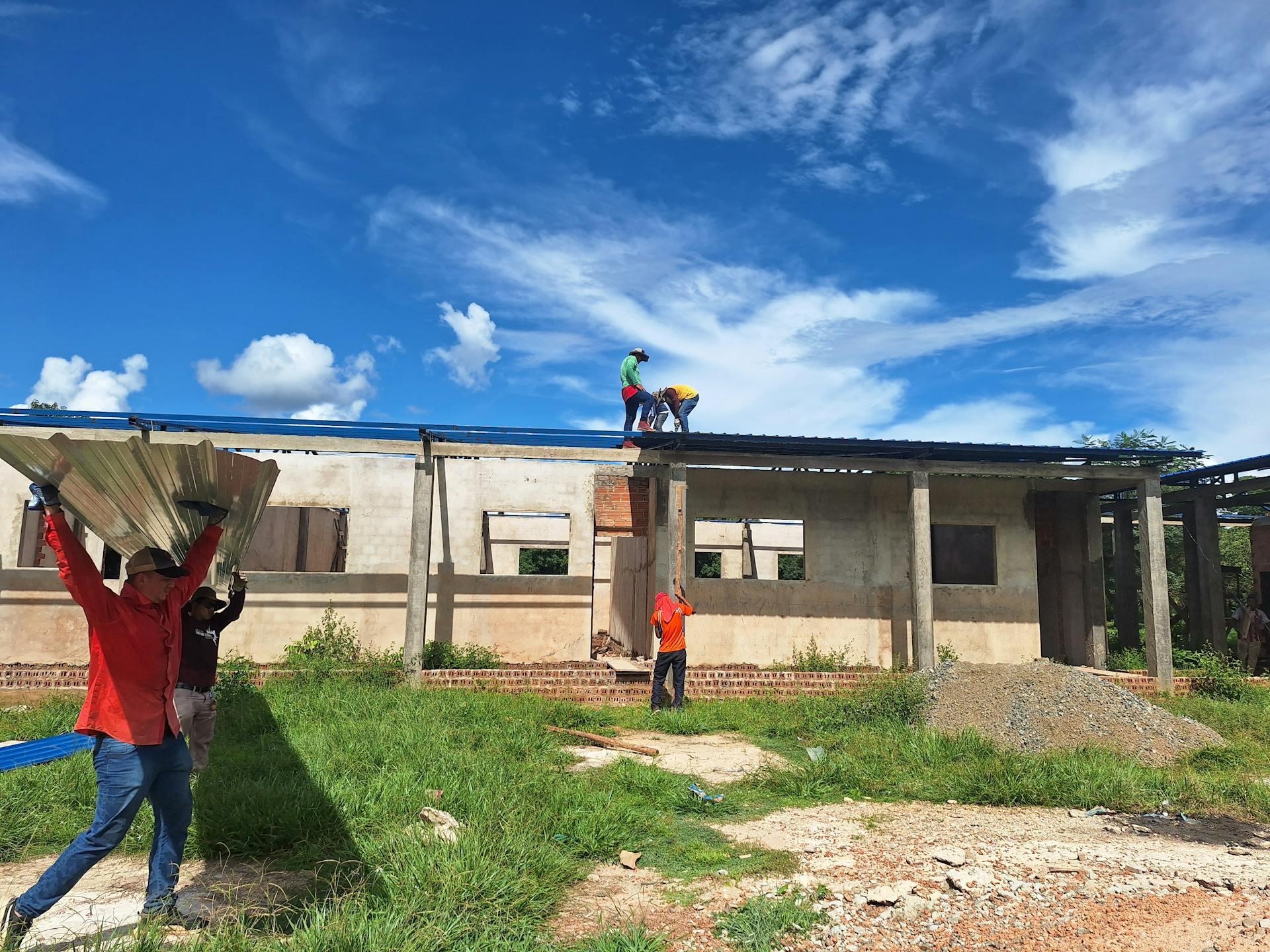 Workers install roofing on an unfinished building under a bright blue sky.