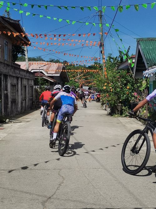 Free Cyclists ride through a village street adorned with colorful flags under a clear blue sky. Stock Photo