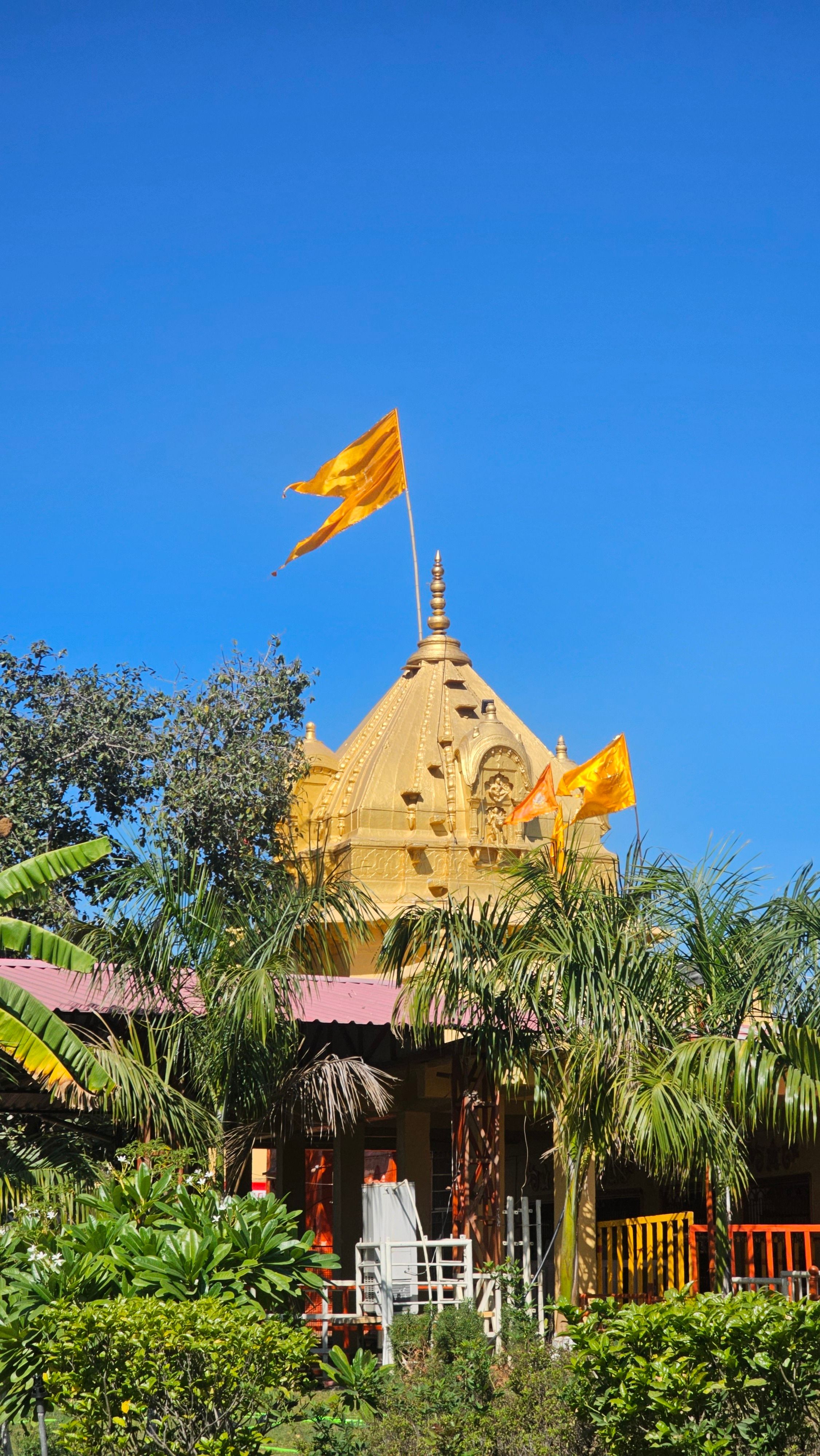 A golden temple with flags rises amidst lush greenery and palm trees, set against a clear blue sky.