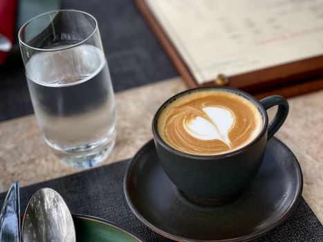 Close-up of a latte with heart-shaped latte art beside a glass of water on a cafe table.