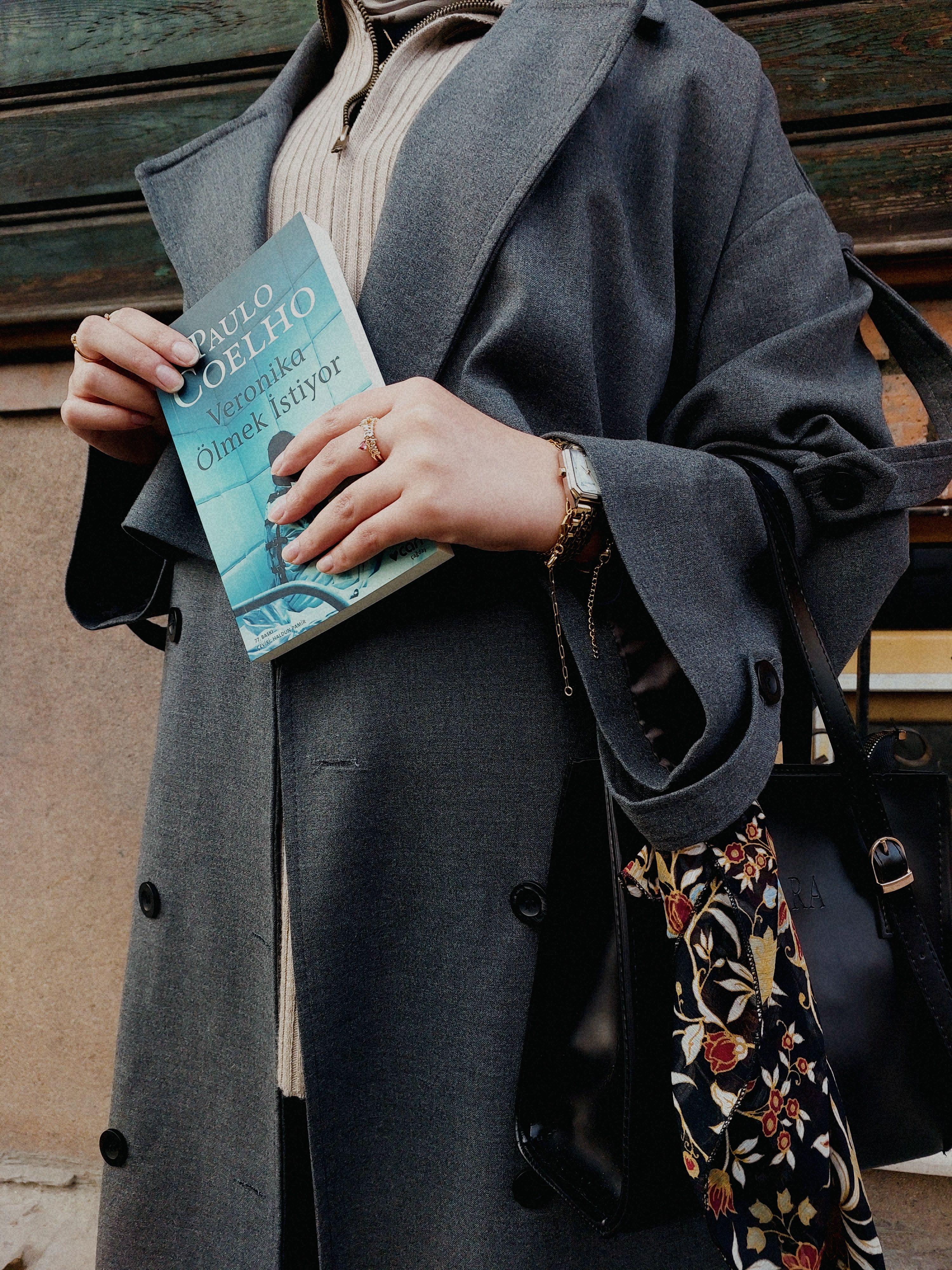 A person holding a Paulo Coelho book, dressed in a gray coat and holding a floral bag.