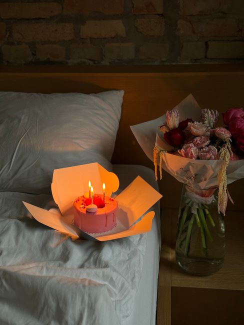 A romantic indoor scene with a lit birthday cake and bouquet on a bedside table.